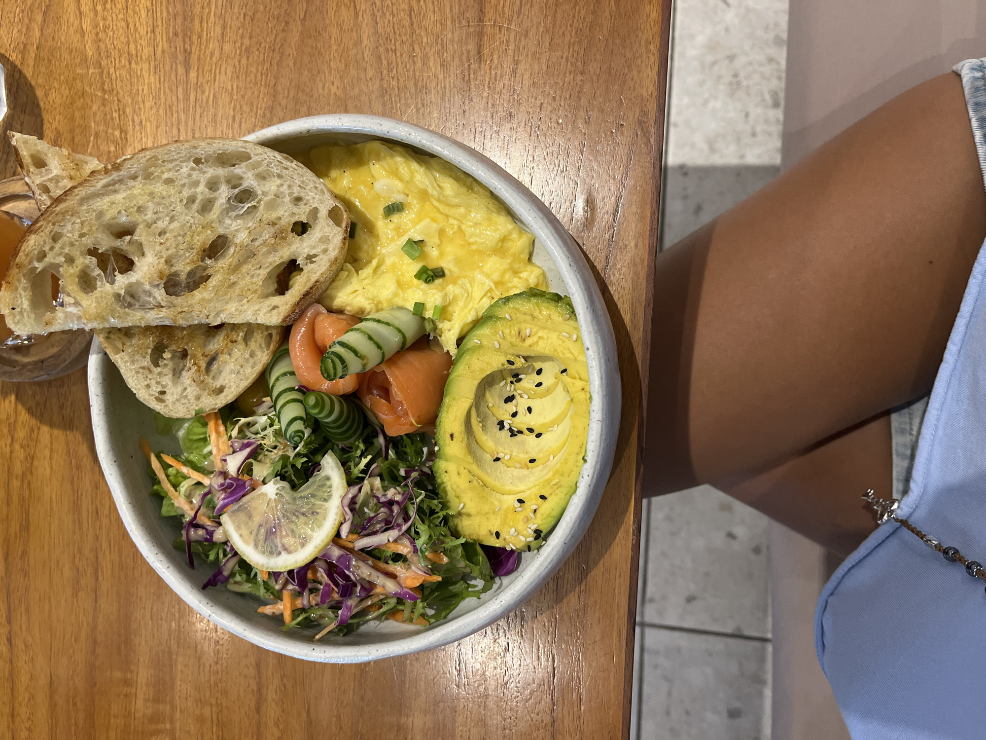A close-up, overhead shot presents a bowl of food placed on a wooden table. The bowl contains scrambled eggs, toast, avocado, salmon, cucumber, and a salad with a lemon slice. The salad appears to be mixed greens with shredded red cabbage and carrots. Black and white sesame seeds garnish the avocado. Next to the table, a person wearing a light blue top is visible. The setting appears to be a cafe or restaurant. Given the cuisine style and the overall ambiance, it is possible the image was taken in a location similar to establishments found in the city of Kuta Selatan, Indonesia, but no concrete evidence is provided to ascertain.