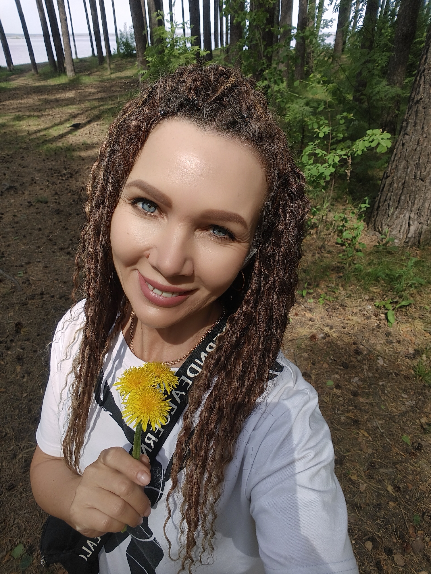The image depicts a woman with light skin, blue eyes, and brown, braided hair. She is smiling and holding a small bunch of yellow dandelions. She is wearing a white t-shirt and a black strap with white lettering is visible across her shoulder. The background consists of a forest with tall, thin trees and scattered foliage. The ground is covered in brown leaves and needles. In the distance, water can be seen between the trees. The location is in a forest, possibly near a body of water. The city and state are not specified.