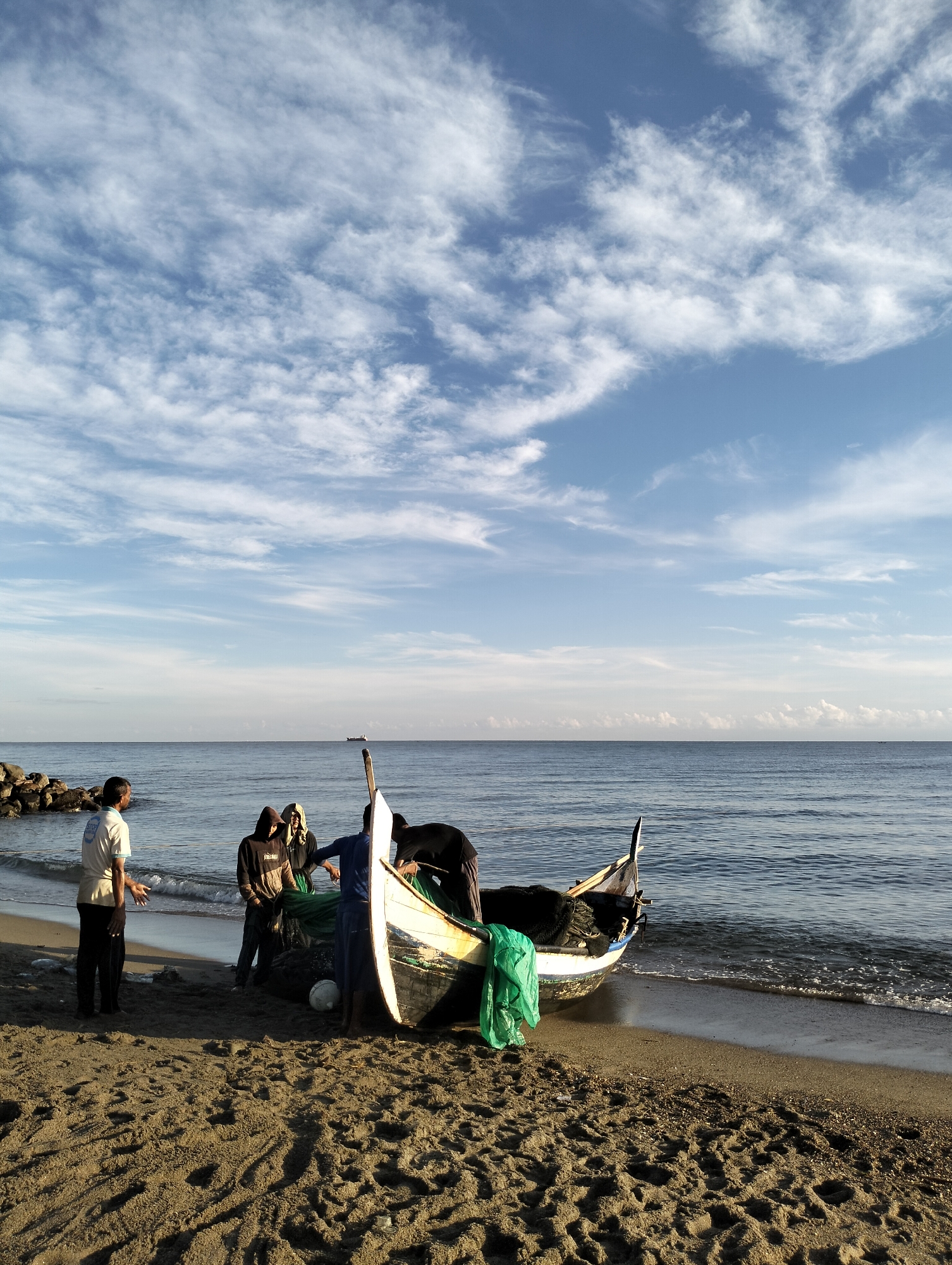 The image shows a group of people gathered around a small fishing boat on a beach in Lhokseumawe, Indonesia. The boat is partially on the sand and partially in the water, with fishing nets and a green plastic bag inside. There are 5 people. One person stands off to the side and appears to be talking or gesturing towards the boat. Three other people are working around the boat. One other person is in the boat. The beach is sandy with distinct footprints. In the background, there is the ocean with gentle waves. The sky is blue with scattered white clouds. A line of rocks is to the left. In the distance, there is a ship.