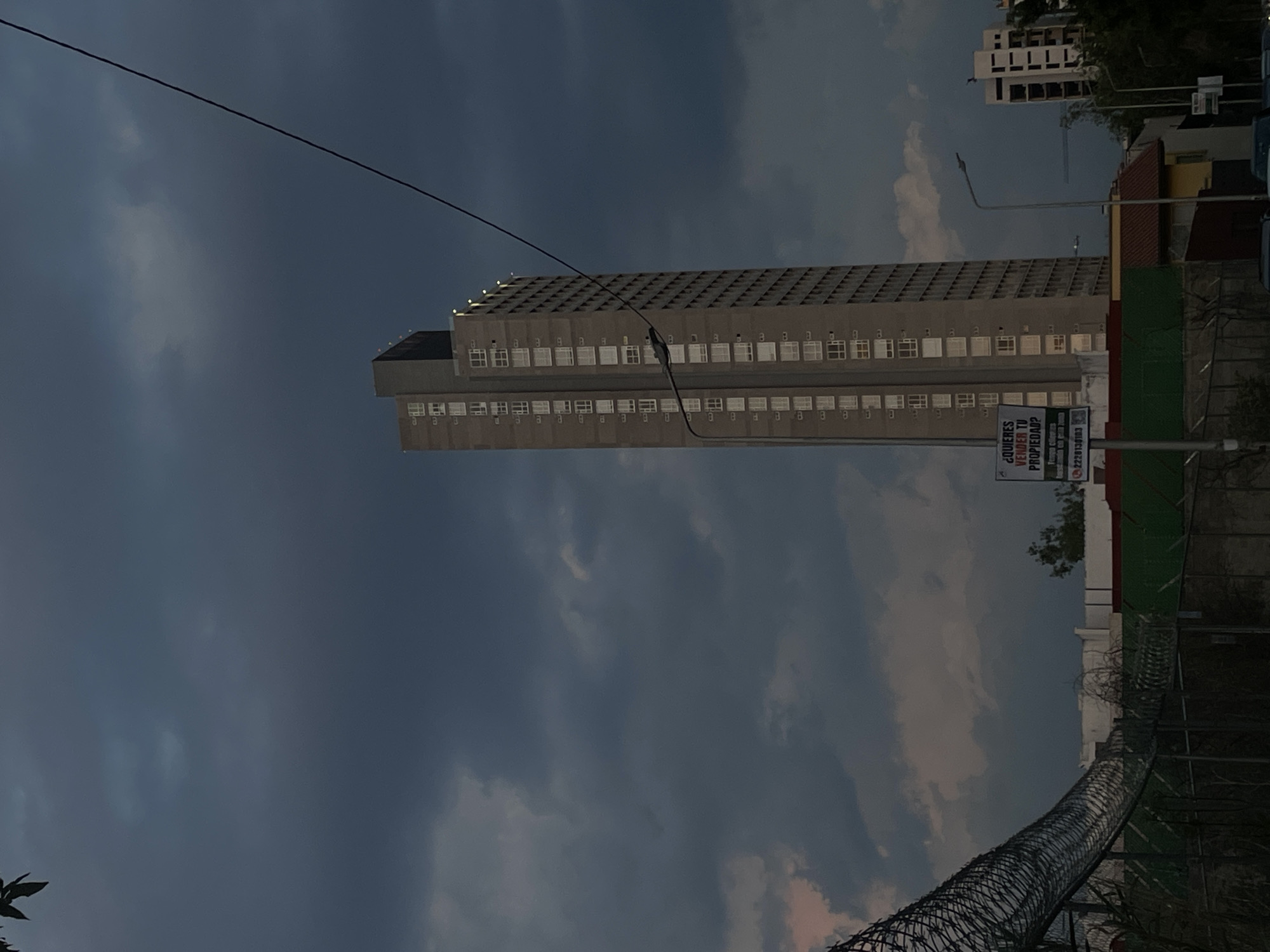 The image shows a tall, grey concrete building with multiple rows of windows, likely an apartment complex or office building, set against a cloudy sky. A sign on the right side of the building promotes property sales with a phone number listed. A barbed wire fence runs along the bottom right portion of the image. A street light is visible on the right. The building is the Torre de Agua Azul, a landmark in Puebla, Mexico.