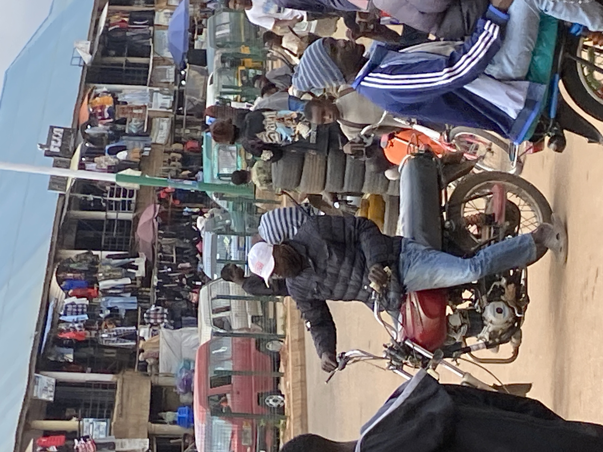 This image captures a bustling street market scene in Bukuru, Nigeria, filled with people, vehicles, and various goods for sale. The setting is an outdoor, possibly unpaved, street lined with numerous market stalls and multi-story buildings housing shops.
In the foreground, a person wearing a black puffer jacket, blue jeans, and a white baseball cap is seated on a red motorcycle, holding the handlebars. Further back, another person on a motorcycle is visible, wearing a blue and white striped top and a blue and grey track jacket with white stripes. Many other individuals are scattered throughout the market, some walking, some gathered around stalls, and several more on or near motorcycles, suggesting active commerce and transportation.
The primary activity depicted is daily market life, with vendors selling goods and people commuting or shopping. Market stalls, some covered by large umbrellas (blue and reddish-brown), display a variety of items, including clothing like shirts, trousers, and dresses, hanging on racks and laid out on tables. A prominent stack of vehicle tires is visible in the mid-ground. Several vehicles, including a red mini-bus or van and other lighter-colored vans, navigate or are parked along the busy street.
The time of day is bright daytime, indicated by the clear blue sky and strong illumination. The overall atmosphere is busy and active. A notable detail is a sign affixed to a building, clearly displaying the text "USA 979".