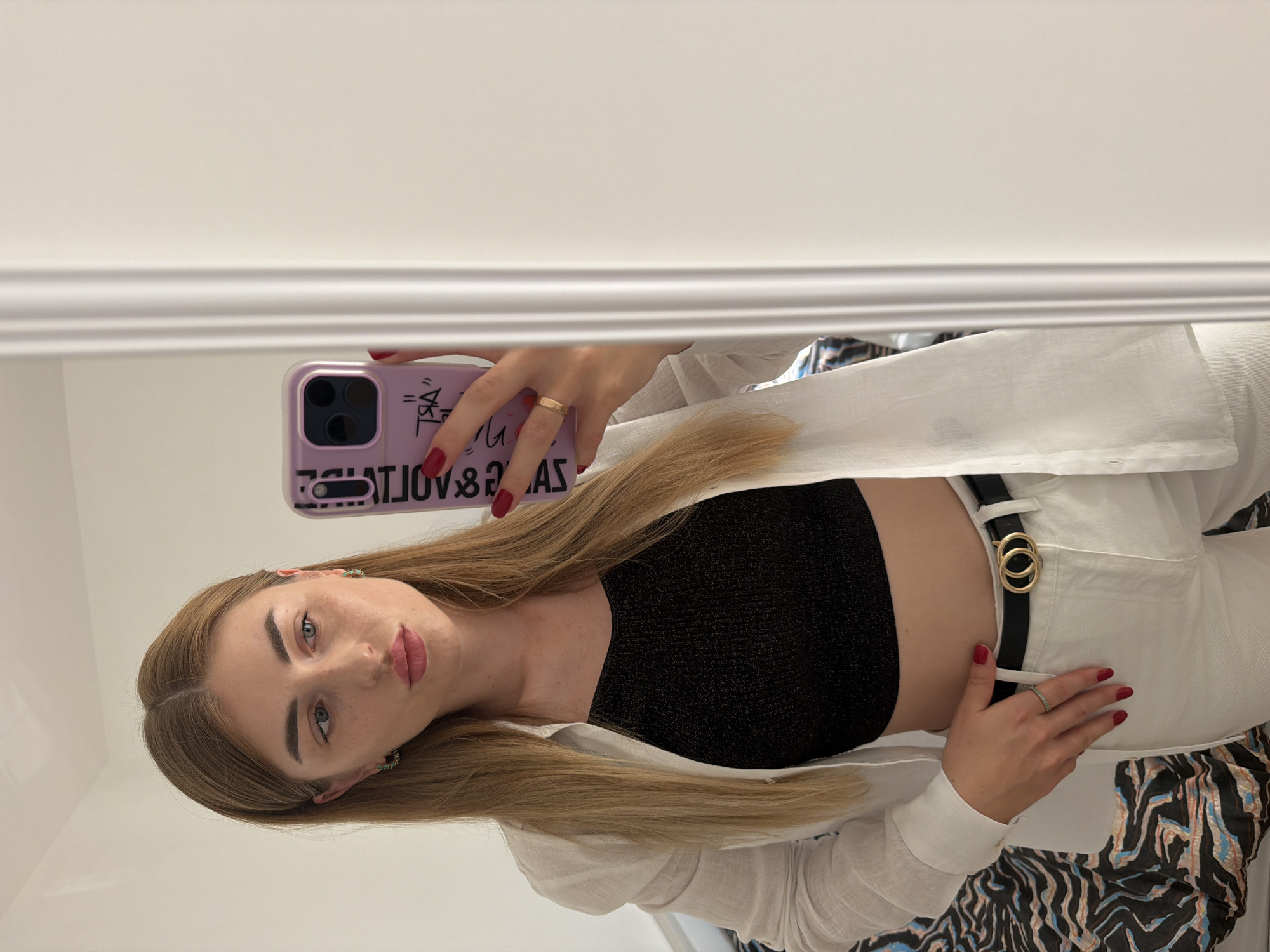 The media file shows a young woman taking a mirror selfie. She has blonde hair and is wearing a white open shirt over a black crop top and white pants. A black belt with a gold double-ring buckle cinches her waist. She is wearing a gold ring on her right ring finger and teal earrings. Her nails are painted red. The background is a plain, light-colored wall with decorative molding. The lighting appears to be indoor and even. The woman is looking directly at the camera with a neutral expression. Her phone, which has a purple case with black text, is visible as she holds it up to take the picture. The text on the phone case is reversed and appears to read "JUST A VOLTAGE". The image is oriented vertically, suggesting it was taken with the phone in portrait mode.