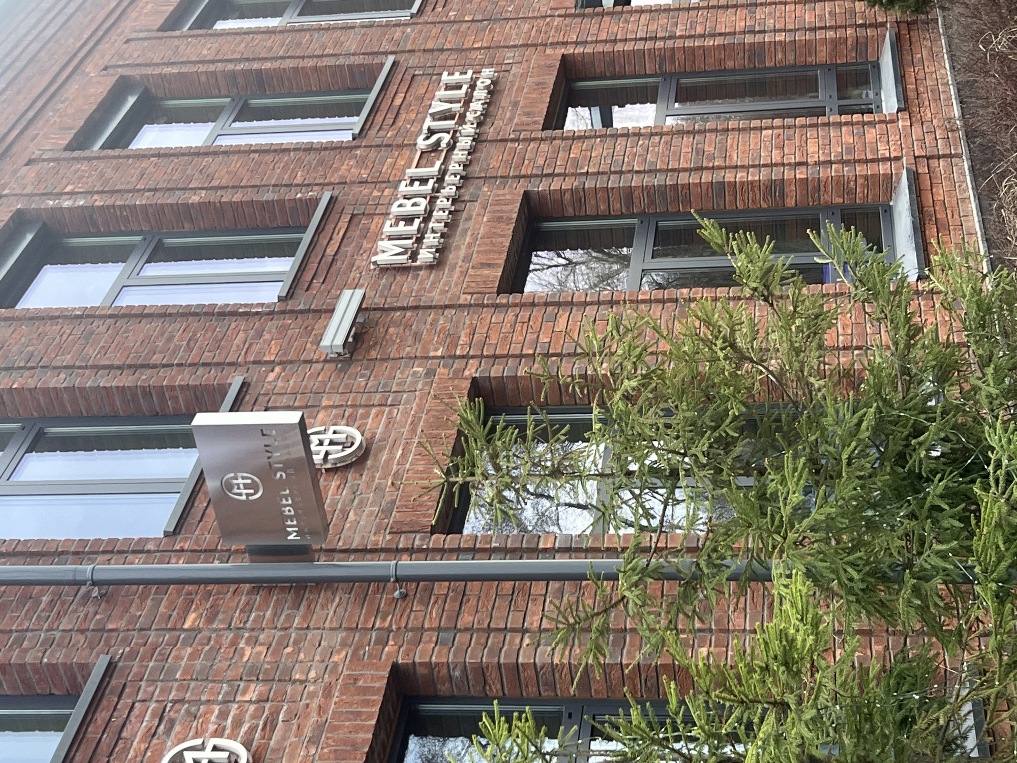 The image displays a low-angle, upward view of a multi-story building constructed with distinctive red-brown brickwork, located in Kazan, Russia. The architecture features deeply set windows with dark grey frames and horizontal brick banding that separates the floors.
Prominently displayed on the building's facade are signs for a business called "MEBEL STYLE." A large vertical sign in the upper-middle section reads "MEBEL STYLE" in white capital letters, with "ИНТЕРЬЕРНЫЕ САЛОНЫ" (meaning "INTERIOR SALONS" in Russian) in smaller white text beneath it. Below this, a silver-grey rectangular sign juts out from the wall, featuring a circular logo resembling a stylized "H" or interwoven pattern, alongside "MEBEL STYLE" written vertically. A standalone circular logo, identical to the one on the projecting sign, is also visible on the brick wall below it.
A dense, green evergreen tree, possibly a spruce or fir, occupies the lower right and central foreground, with its branches partially obscuring the building's windows and brick facade. The windows reflect the bright, possibly overcast, daytime sky. A grey horizontal pipe or gutter system runs across the mid-left portion of the building. The scene is static, with no visible human activity.