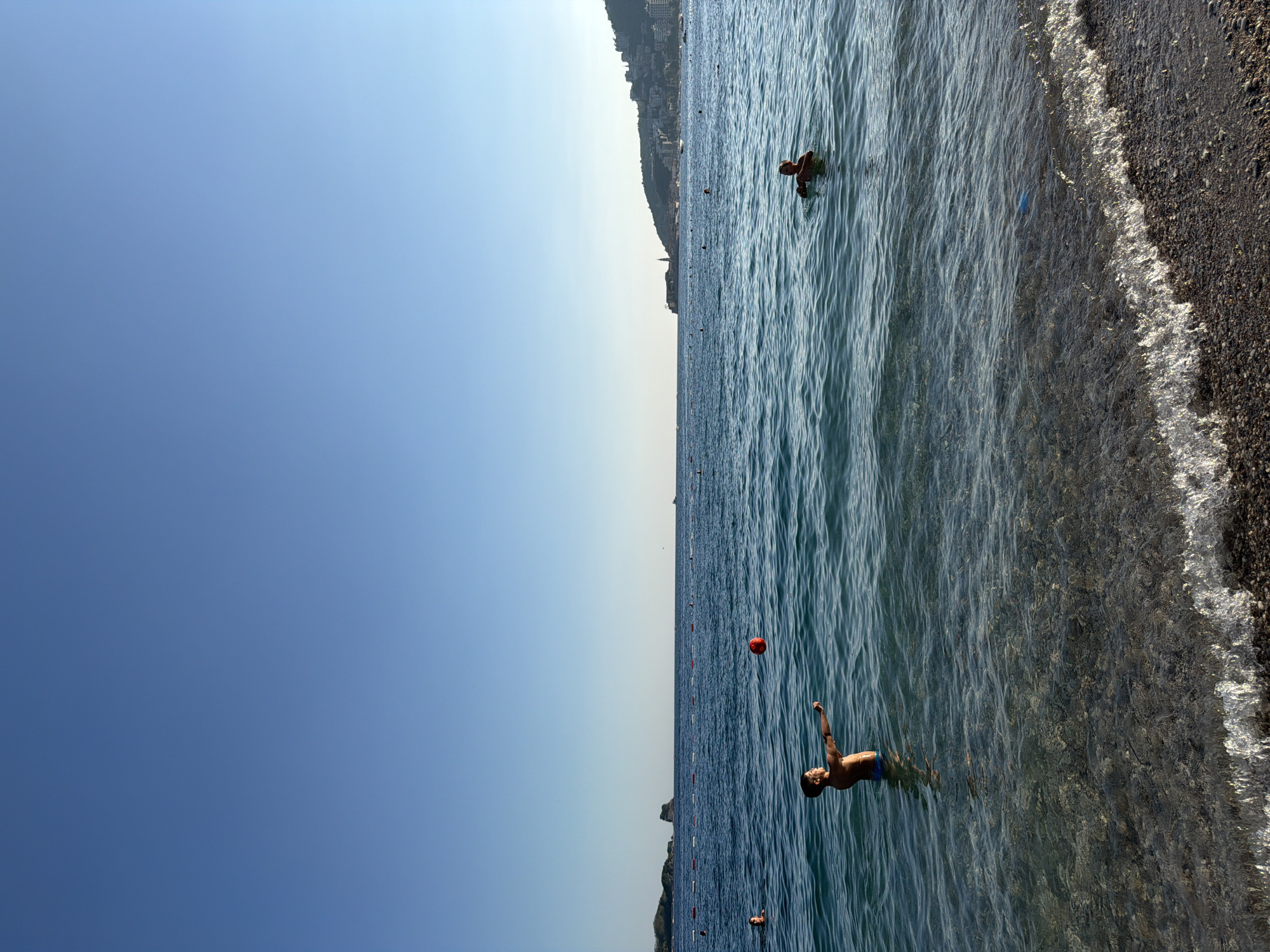 The image depicts a sunny day at a beach in Budva, Montenegro. The scene captures a body of water with two individuals visible. In the foreground, a young boy stands in the clear, shallow water, with his body turned to the left and his right arm extended, as if playing with a ball. A bright red ball is suspended in the air in front of him. In the mid-ground, another person is partially submerged in the water, their head and shoulders visible. The water is calm and has visible pebbles and rocks on the seabed. The background shows the coastline of Budva, with buildings dotting the hills and a clear blue sky overhead. A line of buoys stretches across the water, marking a boundary. The overall impression is one of a serene beach day.