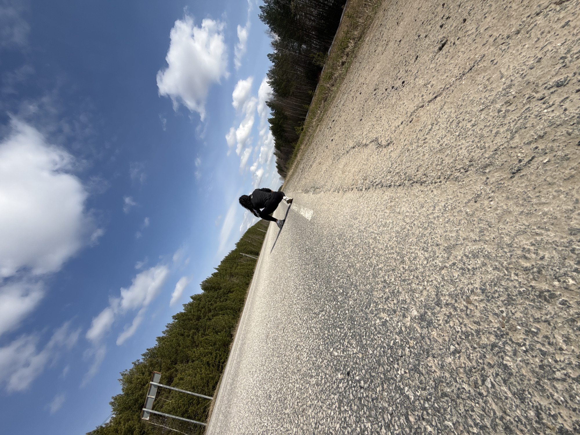 A low-angle shot presents an unidentifiable individual in black clothing, crouched on a concrete surface marked with a white line, ostensibly a rural road in the vicinity of Maksimovo, Russia. The person's orientation suggests possible interaction with the road's surface. Adjacent to the road are dense forests bordering the scene on either side. Above, a bright blue sky is dotted with scattered white clouds. The location is indicated by the image's origin and metadata.