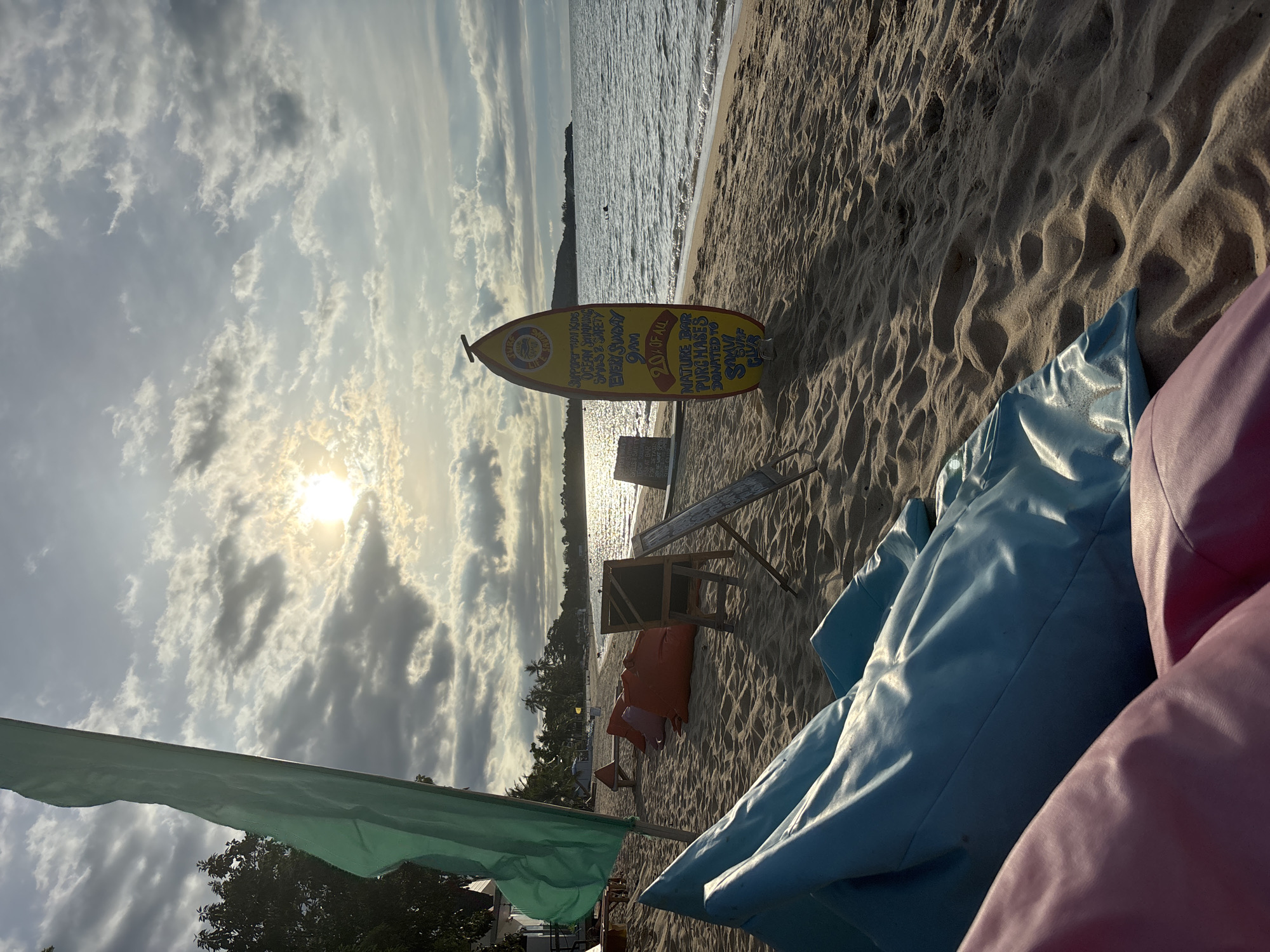 The image depicts a beach scene in Baan Mae Nam, Thailand. A surfboard with the text "SUPPORT THAI KIDS OCEAN SHIMMING SKUS & SAFETY EVERY SUNDAY 9AM 20% OF ALL NATURE BAR PURCHASES DONATED TO S SURF" is situated on the sand near the water's edge. The beach is sandy, with a few lounge chairs and bean bags visible in the foreground. A green flag is visible on the left side of the image. The sky is cloudy, with the sun shining through, reflecting on the water. In the distance, there appears to be a landmass with trees.