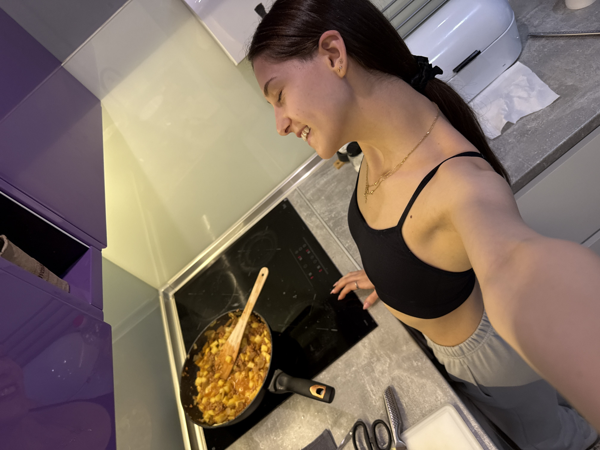 A young woman is cooking in a modern kitchen. The kitchen features purple cabinets on the left, a grey countertop, and a black induction cooktop with a frying pan on it, containing what appears to be a dish made of potatoes and meat. The woman is wearing a black sports bra and grey sweatpants. She is smiling and seems to be enjoying the cooking process. A wooden spoon is in the pan. There are scissors and a cutting board on the counter near the stove. A white appliance is on the countertop in the background. The location is a kitchen in Sofia, Bulgaria.