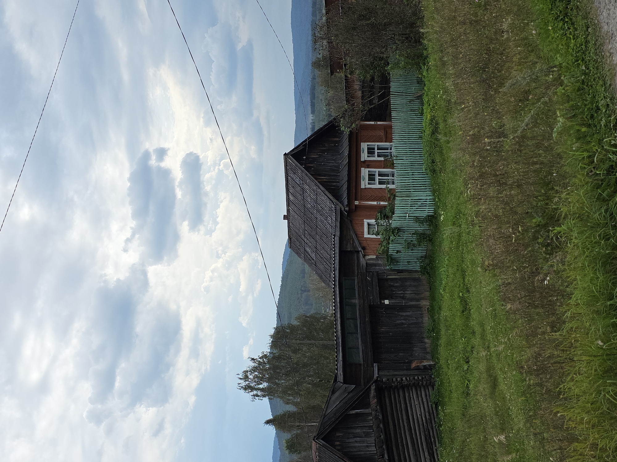 The image depicts a rural scene under a cloudy sky. In the foreground, a grassy field with tall dry grasses and some green vegetation is visible. Behind the field, there are several wooden structures, including a log house on the left and a red-painted house with white-trimmed windows in the center. A light blue-green picket fence encloses a portion of the property in front of the red house. In the background, rolling hills covered in trees and vegetation are visible, with more distant hills fading into a hazy horizon. Power lines are strung across the sky. The scene appears to be in a village setting.