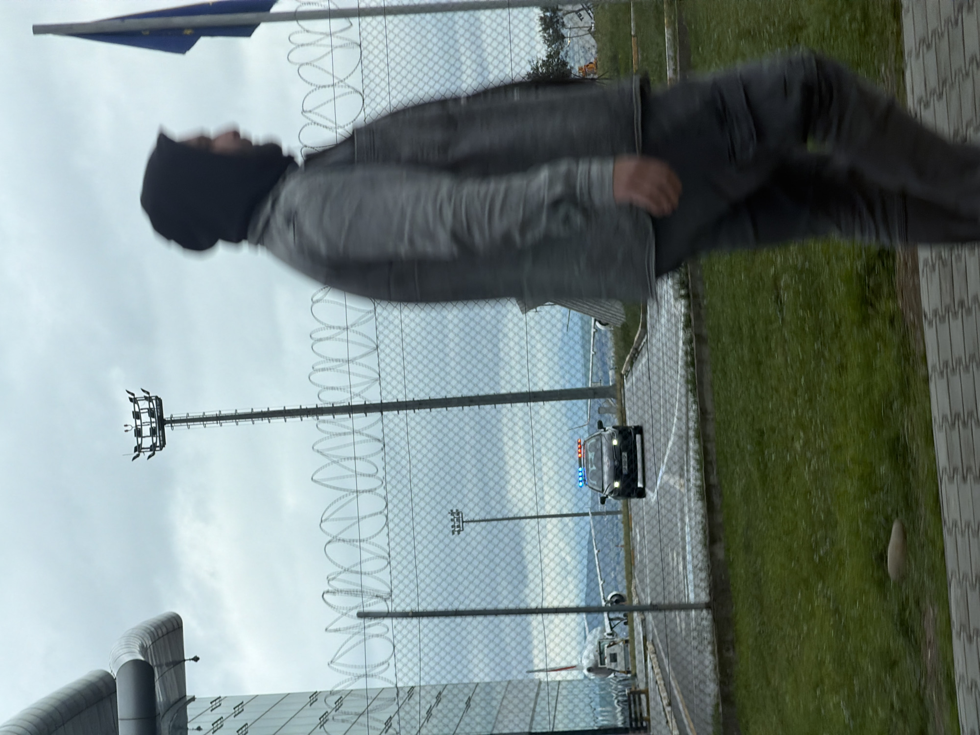 The image depicts a person wearing a black beanie, a grey jacket, and grey pants, positioned in front of a chain-link fence topped with barbed wire. Beyond the fence, a police car with its lights activated is visible on a tarmac, along with parked airplanes. The location is identifiable as the Tbilisi International Airport in Georgia due to the presence of airport infrastructure and the Georgian flag. Additional elements include light poles, green space, and part of an airport building in the foreground.