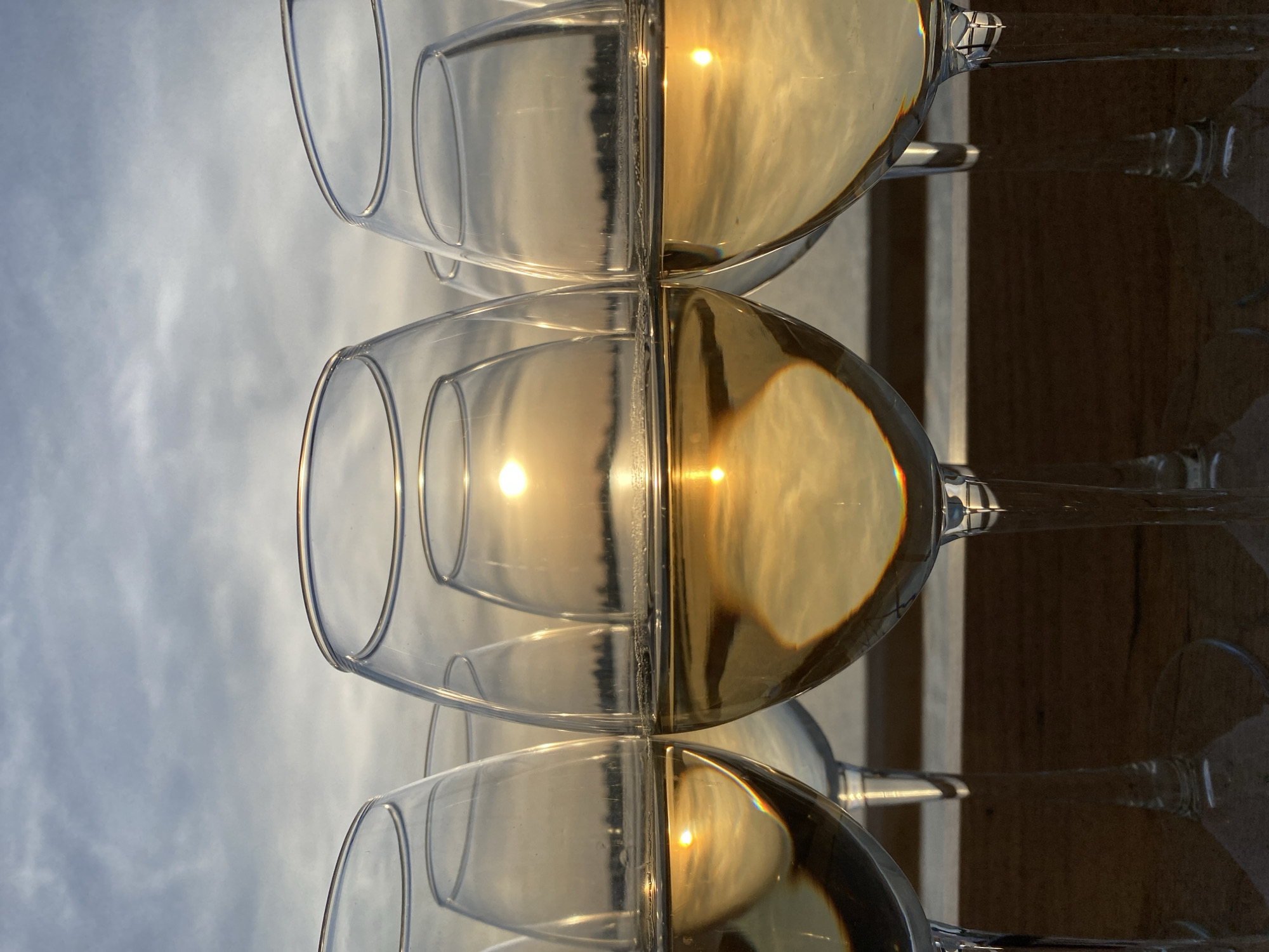 The image depicts a row of wine glasses, some filled with a yellow liquid, likely wine. The glasses are arranged on a surface, possibly a table or ledge, with an outdoor background visible through the clear sections of the glasses. The background appears to be a sky with clouds. The light is bright, suggesting daylight. The reflections in the liquid-filled portions of the glasses suggest a sunset or sunrise scene. The focus is on the glasses, with the background somewhat blurred. There is no indication of this image being located in the city of Aramil, Russia.