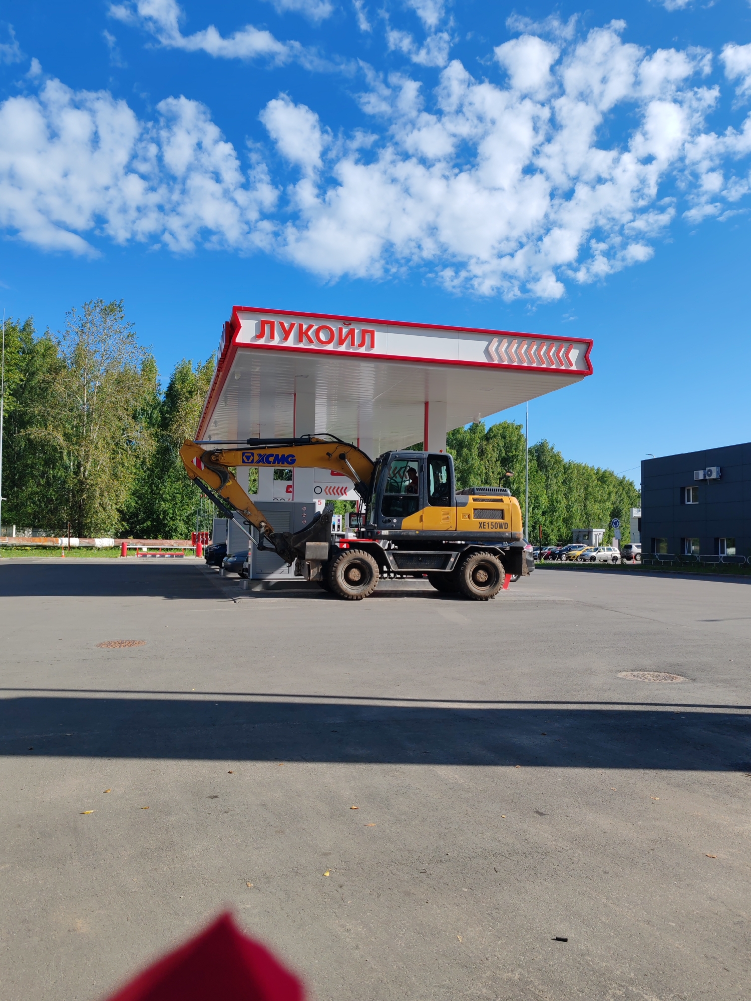 The image shows an excavator parked on the asphalt surface of a Lukoil gas station in Syktyvkar, Russia. The excavator is yellow and black and has the model number "XE150WD" visible. It is positioned beneath the gas station's canopy, which is red and white and features the Lukoil logo. Behind the gas station, a line of trees forms a backdrop, and a dark-colored building with windows is visible to the right. The sky is blue with scattered white clouds. The lighting suggests it is daytime.