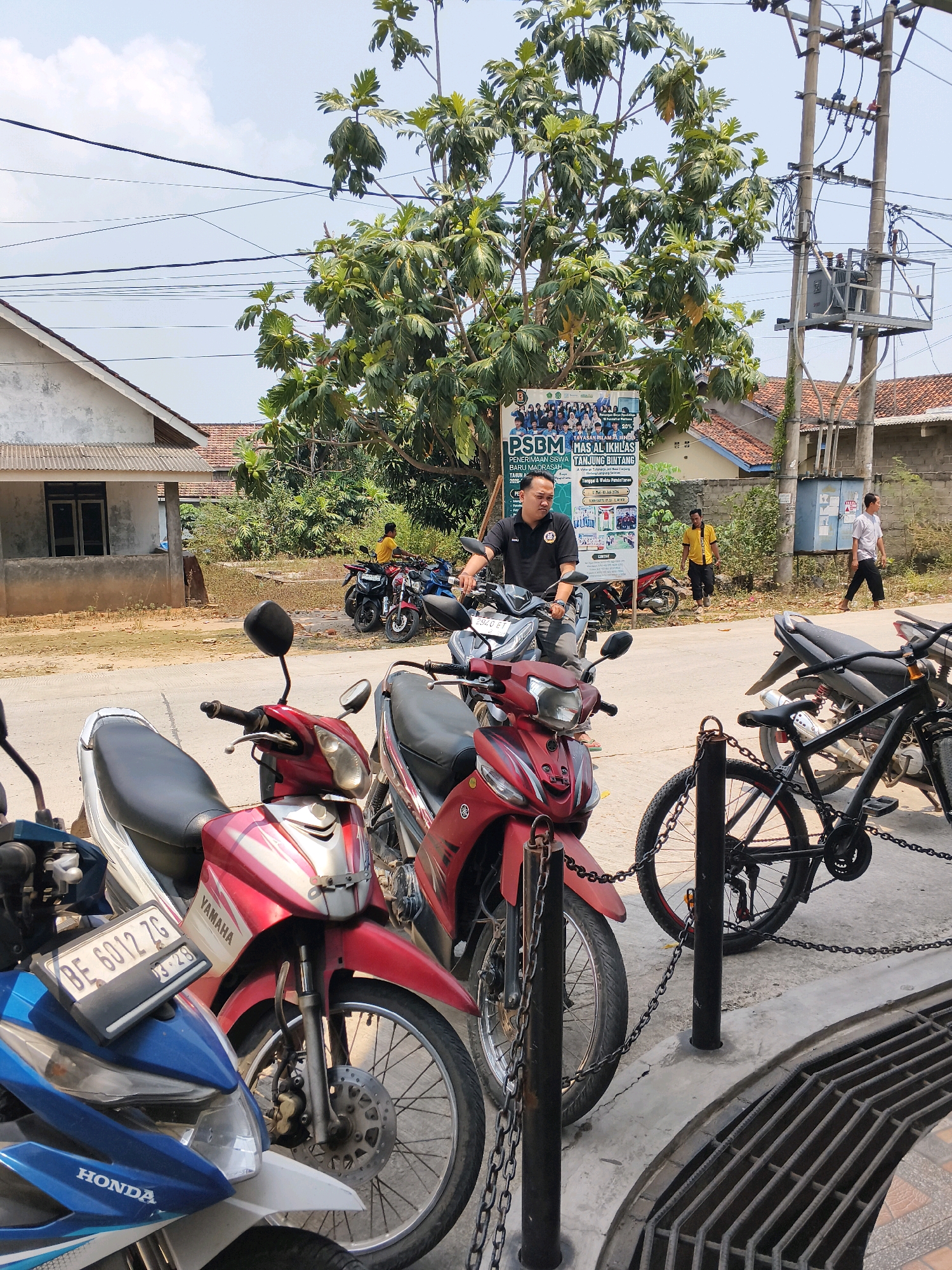 In Sabah Balau, Indonesia, on a sunny day, a man in a black t-shirt and dark pants sits on a red motorcycle, looking towards a billboard behind him. The billboard displays information about PSBM MAS AL IKHLAS BARU MADRASAH TANJUNG BINTANG, featuring a group photo of students and staff. Other motorcycles are parked nearby, including a blue Honda and a black bicycle with a chain. The environment includes a building with a tiled roof, a utility pole with electrical equipment, and dense foliage from a large tree. Two other individuals are visible in the background: one man in a yellow shirt and another in a light-colored shirt walking. The foreground shows a metal grate on the ground and a concrete walkway.