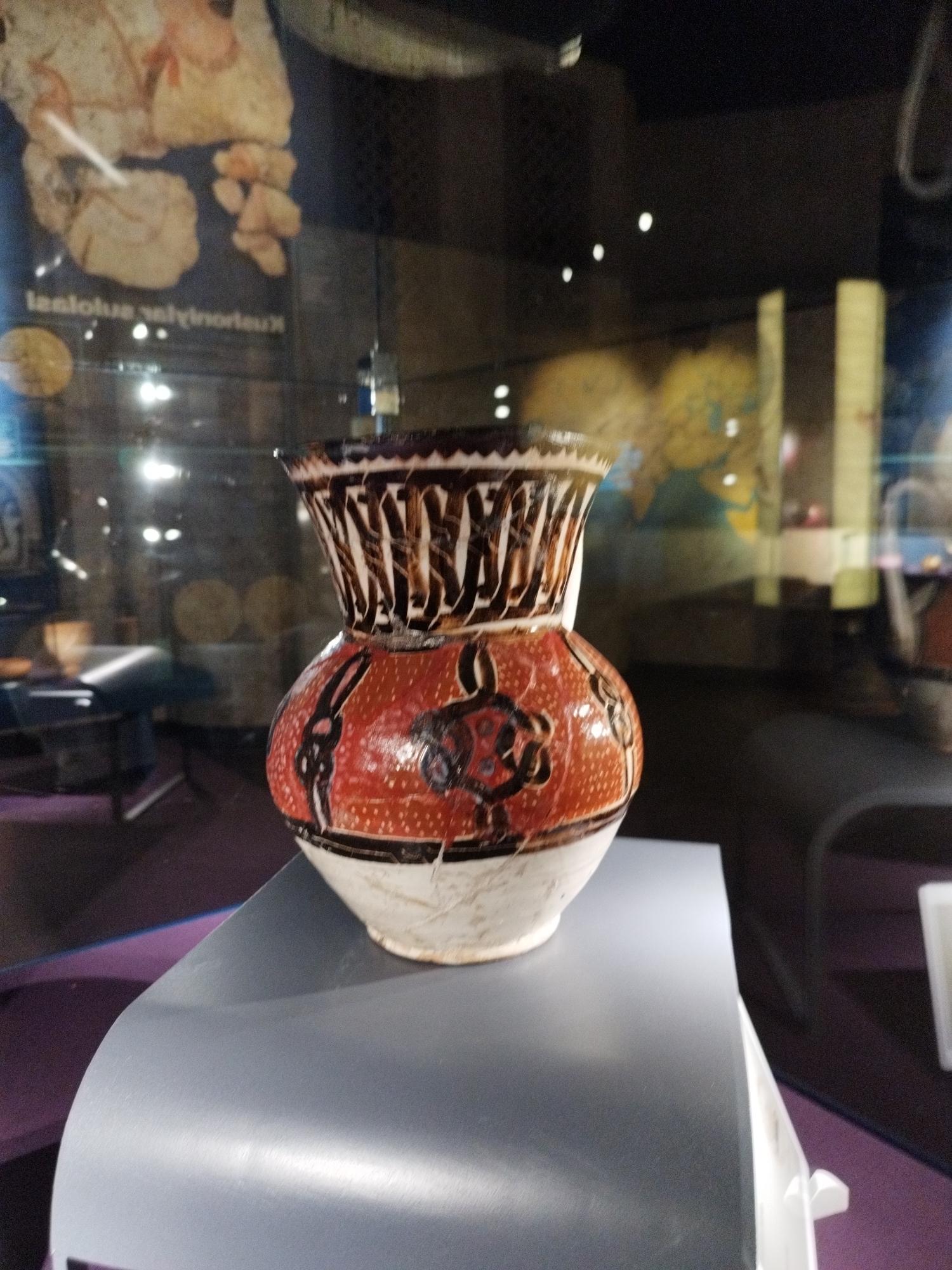 A close-up, eye-level shot shows an ornate pottery vase displayed in a museum. The vase has a white base, a red midsection with black and blue decorative patterns, and a white and brown patterned top. It is presented on a sleek, light gray pedestal. Behind the vase, a glass display case reflects the surrounding environment, showing blurred images of other exhibits and lighting. The background is dark, with some blurry images of what appear to be maps or murals on the wall.