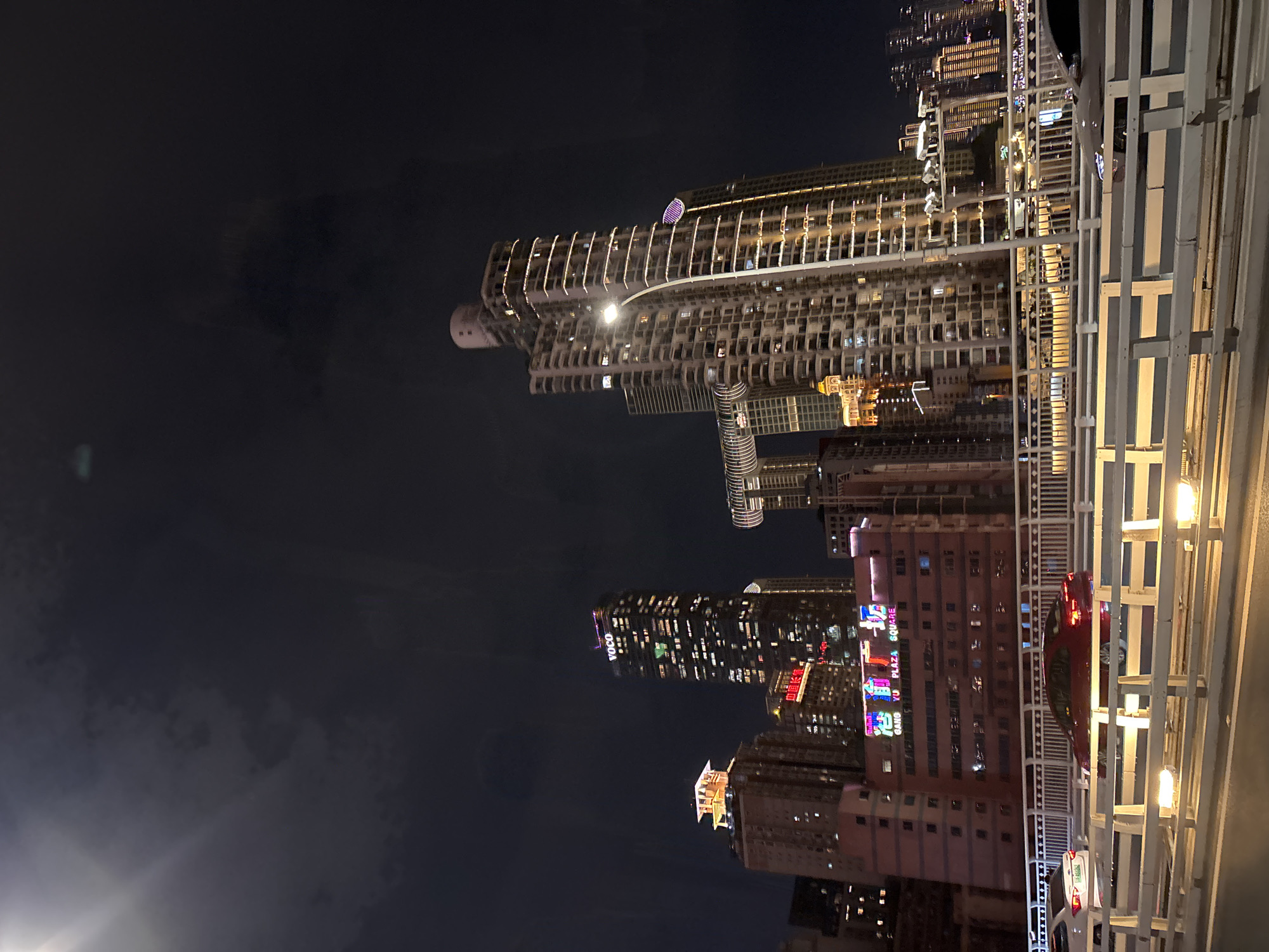 This is a nighttime photograph taken from a bridge overlooking a city skyline in Yuzhong District, China. The sky is dark with some clouds and a bright lens flare in the upper left corner. Numerous tall buildings, illuminated with artificial lights, dominate the scene. One prominent skyscraper, labeled "VOCO" at its apex, is centrally positioned and displays rows of lit windows. To its left, another building has a golden-lit roof structure. To the right of the "VOCO" building, a building with a unique curved architectural feature can be seen. In the foreground, the white metal railing of the bridge stretches across the bottom of the frame, with lights visible along its structure. Cars are seen driving on the bridge, with a red car in the middle lane and a white car in the left lane. A pink building with illuminated signage that reads "GANG YU PLAZA SQUARE" is visible beneath some of the taller buildings. The overall impression is a bustling urban environment at night.