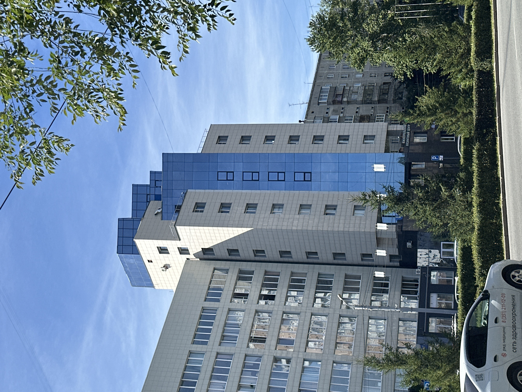 In an urban outdoor setting under a bright, clear blue sky, a multi-story building complex is visible. The building on the left is a light-colored, rectangular structure with numerous windows reflecting the sky and surrounding environment. To its right, a taller, more modern building features a prominent section clad in reflective blue glass, creating a striking contrast with the lighter facade. This blue section is vertically oriented and breaks the otherwise uniform grey stonework of the building. Further to the right, an older, multi-story residential building with visible balconies and a weathered facade is present. Evergreen trees and a manicured hedge line the foreground, obscuring the lower portions of the buildings and the street. A white car is partially visible in the lower left corner, with text and a QR code on its side. Power lines are strung across the upper portion of the sky, and a street light is positioned in front of the light-colored building. The overall scene depicts a typical urban streetscape with varied architectural styles.