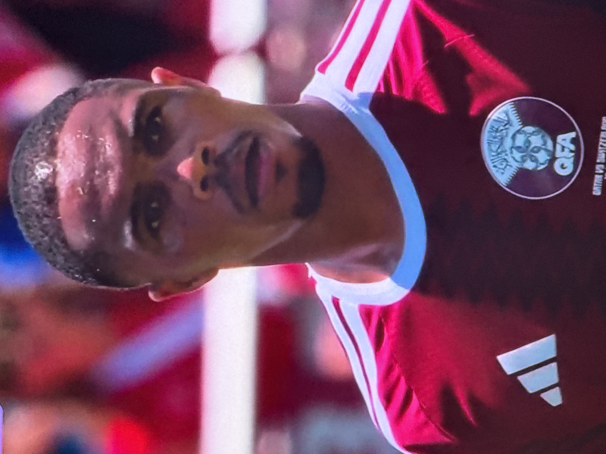 A male soccer player with dark skin and short hair is shown from the chest up, facing forward with his head slightly tilted and his mouth open. He is wearing a maroon jersey with white stripes on the shoulders and a white collar. A white Adidas logo is visible on the left side of his chest, and a circular crest with Arabic text and a soccer ball is on the right side. The background is blurred and appears to be a crowd in a stadium, with some red and pink hues. The lighting suggests an outdoor event during the day. The text at the bottom of the image, partially visible, indicates "QATAR VS SWITZERLAND."