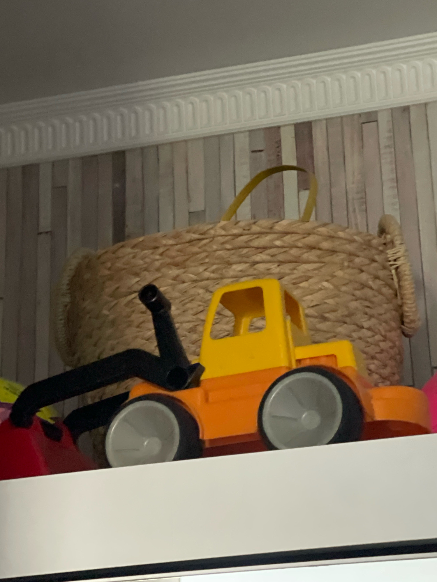 An orange and yellow toy truck sits on a white surface in front of a woven basket and patterned wallpaper. The truck has black and gray wheels and a black arm attached to its front. The basket is large and round with yellow straps. The wallpaper has a vertical striped pattern in shades of light pink and gray. The image is taken from a slightly low angle, looking up towards the truck and basket. There are no people visible in the image.