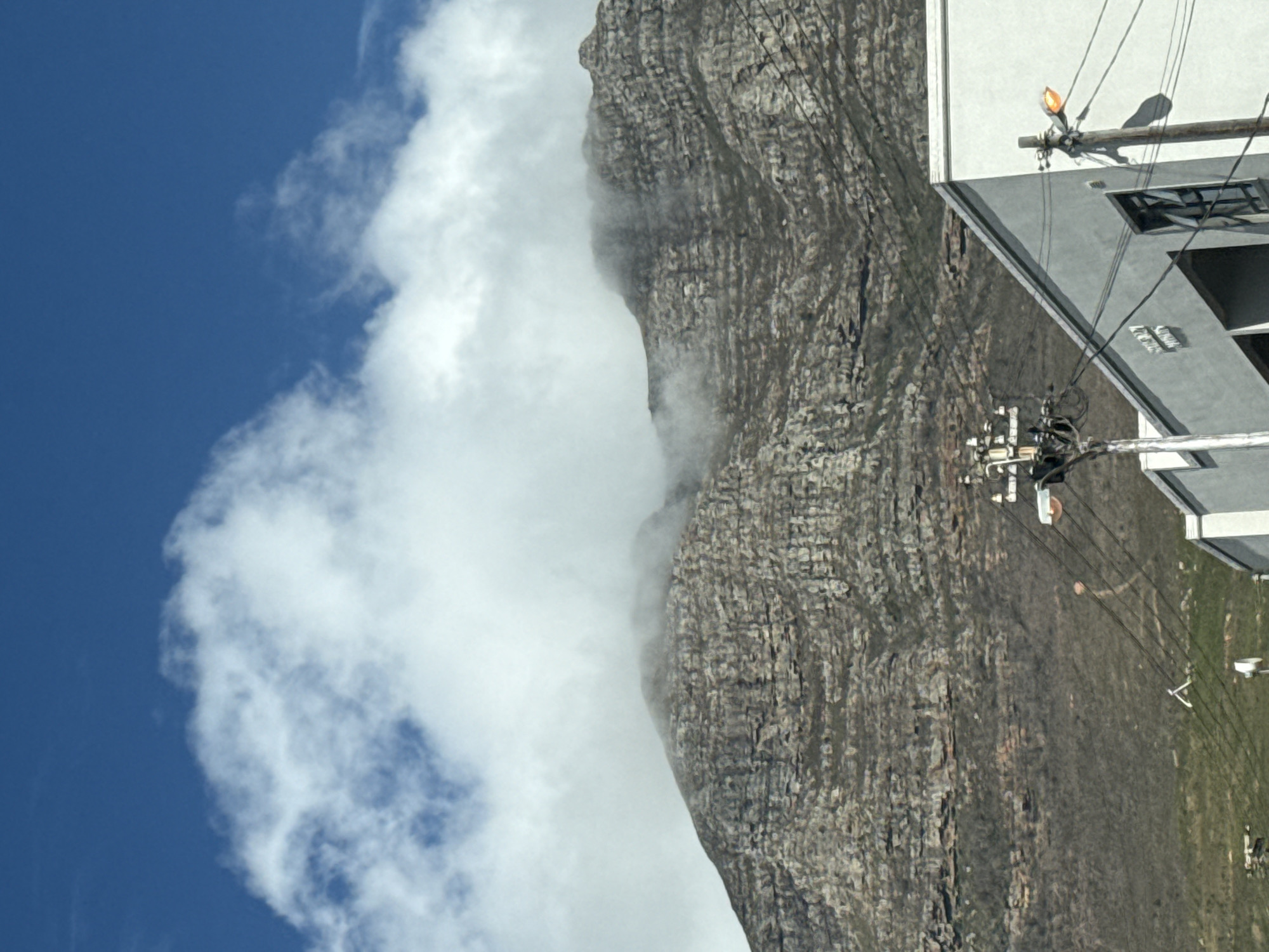 The image captures a view of Table Mountain in Cape Town, South Africa. The mountain's rocky face is partially obscured by a bank of white clouds. Clear blue sky is on the left of the mountain. Utility poles with wires and some parts of a building are to the right. The base of the mountain has a grassy area.