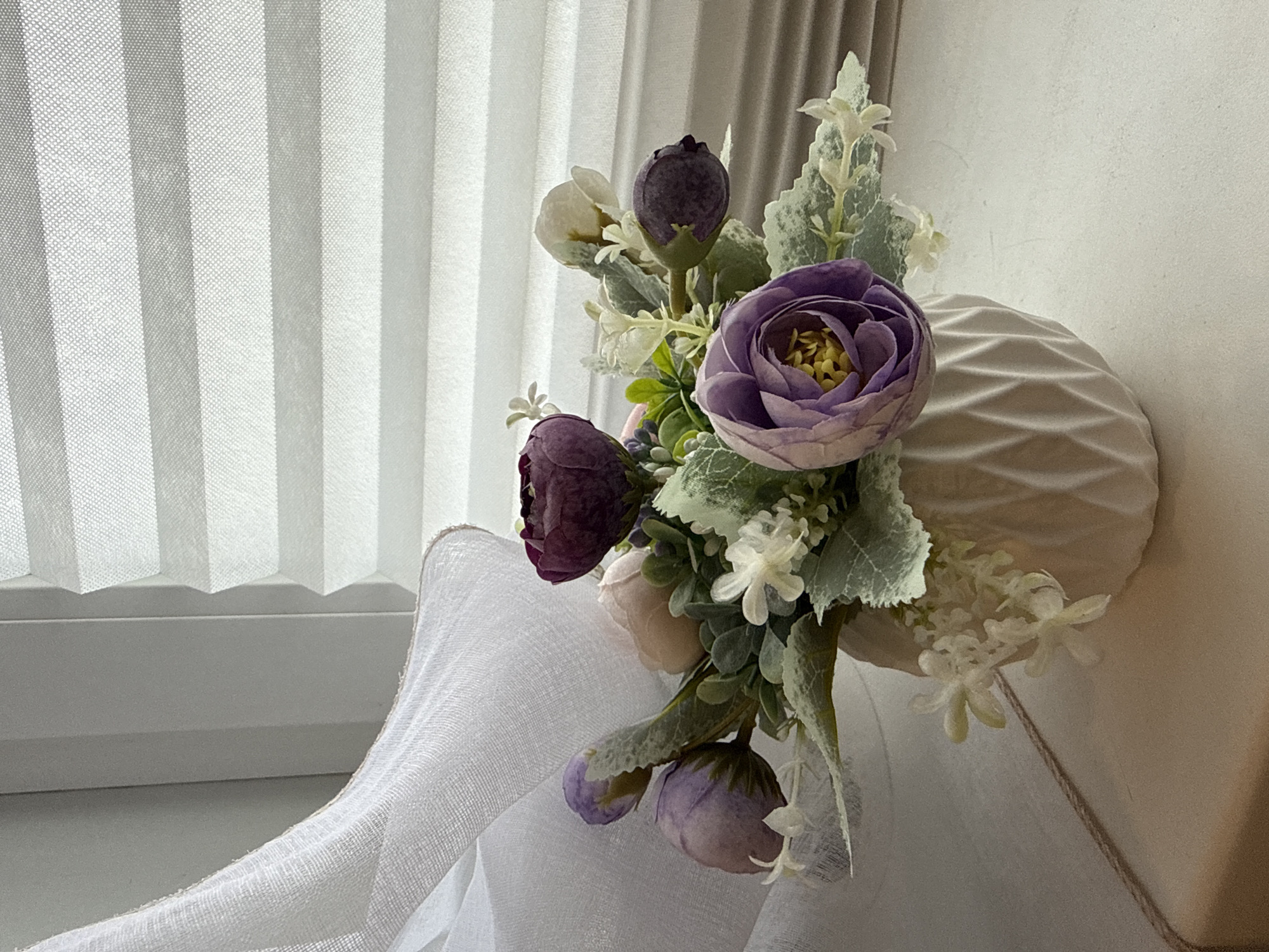 A still life photograph shows a bouquet of artificial flowers in shades of purple and white, arranged in a textured white vase. The vase is positioned on a surface that appears to be a windowsill, with a white pleated curtain in the background and a draped white fabric in the foreground. The lighting suggests it could be daytime, with natural light coming from the left, illuminating the scene. The overall mood is calm and decorative. There is no visible text in the image.