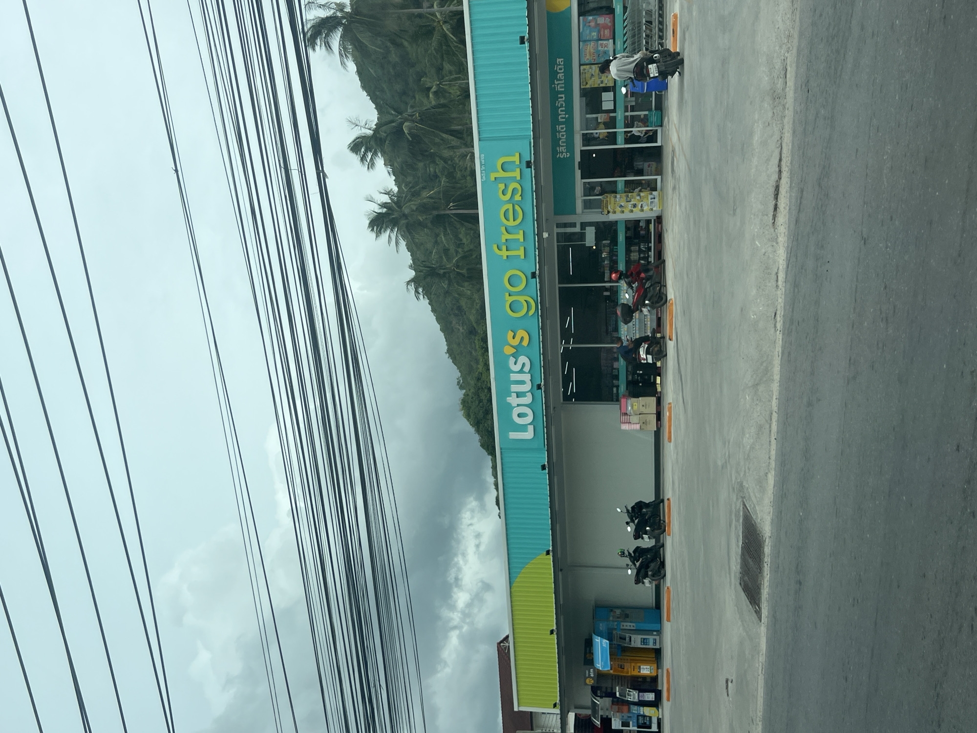 The image shows the exterior of a Lotus's Go Fresh supermarket. The building has a bright teal and green facade with the store's name prominently displayed in white lettering. In front of the store, there are several ATMs and parking spots marked with orange bollards. Two motorcycles are parked in front of the entrance, and a person on a motorcycle is visible near the right side of the frame. Inside the store, shelves stocked with goods are visible through the glass doors, along with shopping carts. The supermarket is situated on a street with a gray asphalt road in the foreground. A hillside covered with lush green vegetation and palm trees rises behind the building, under a cloudy sky. Overhead, a dense network of electrical wires crisscrosses the sky, obscuring a portion of the scene. The overall setting appears to be a tropical or subtropical location, given the presence of palm trees and the type of vegetation.