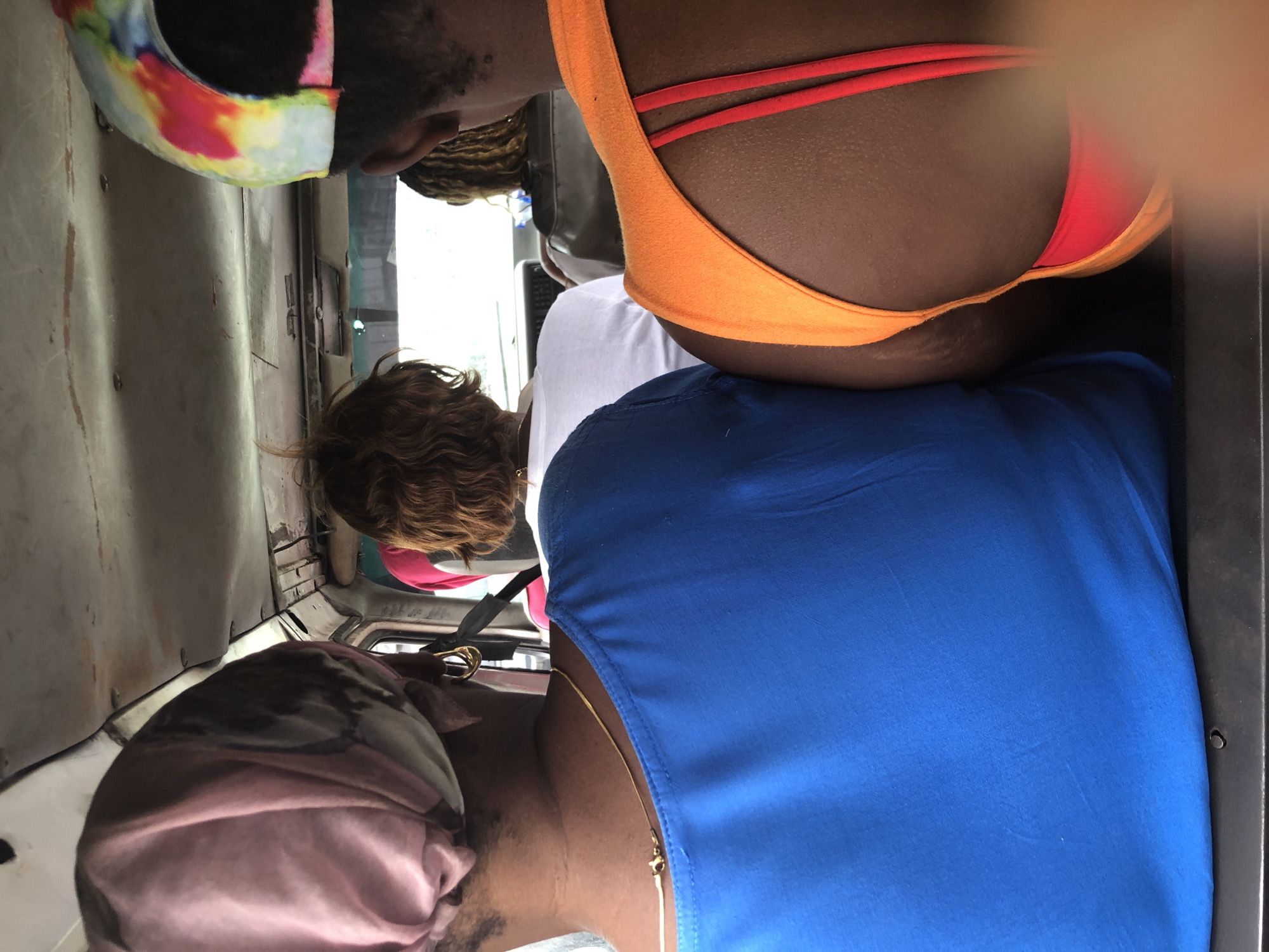 The image captures the interior of a moving vehicle, possibly a bus or van, in Igboefon, Nigeria. The focus is on the rear view of three individuals seated inside. The person in the foreground on the left wears a vibrant blue top and a pink headscarf. To their right, a person with curly brown hair is visible, wearing a white top. The person on the far right is partially visible, wearing an orange top with red straps and a colorful tie-dye cap. The environment appears somewhat worn, with visible ceiling panels and a dashboard. The lighting suggests it is daytime, with natural light filtering into the vehicle. The overall scene depicts a moment of transit within a local setting.