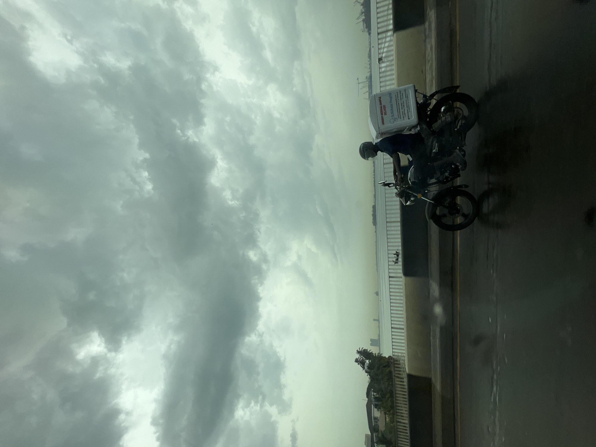 A medical sample delivery driver, likely in Ikoyi, Nigeria, rides a small motorcycle on a road beside a bridge or elevated highway. The driver wears a helmet and carries a white container marked "URGENT MEDICAL SAMPLE DELIVERY" with the words "St Nicholas Hospital" visible. The background features a cloudy sky and a cityscape or waterfront view. The road appears wet or damp.