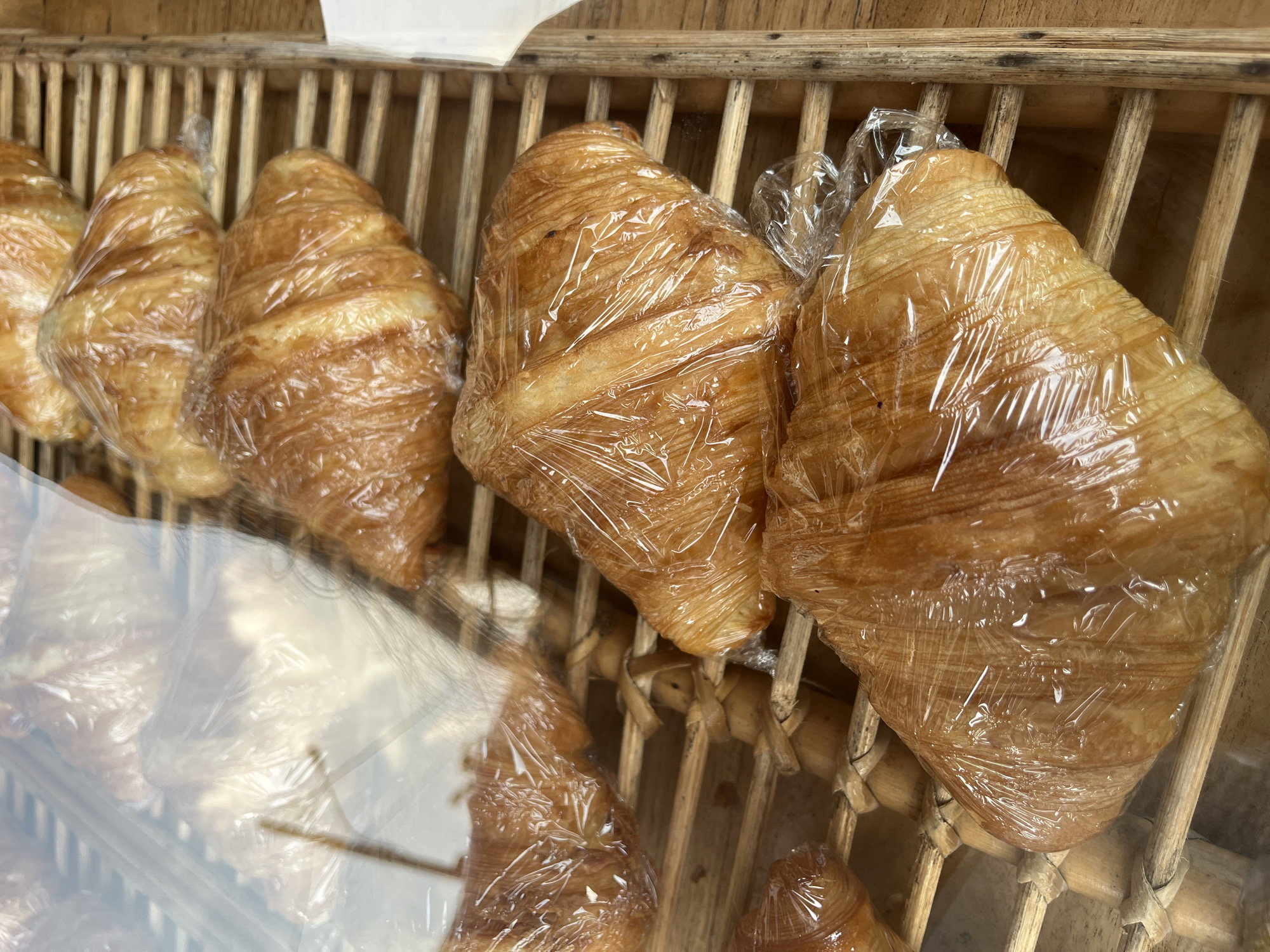 The image depicts a display of croissants in a bakery, possibly in Baturiti, Indonesia, though this is inferred. The croissants are arranged on a light brown, wooden, slatted shelf or rack. Some of the croissants are individually wrapped in clear plastic wrap. The croissants appear golden-brown and freshly baked. The scene is shot at a high angle, looking down into the display. The lower-left corner of the image has some blurry reflections, potentially from a glass display case.