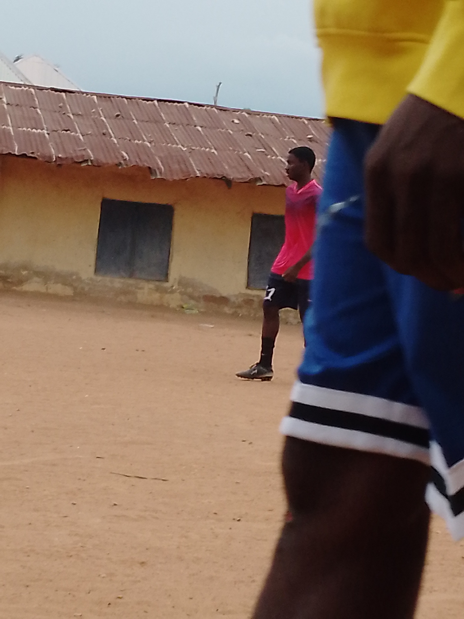 The image shows a dusty open area in Akwanga, Nigeria. A man in a pink shirt and black shorts with the number "7" on it is walking near a small building with a rusty corrugated metal roof and closed windows. In the foreground, the arm and shoulder of another person wearing a yellow shirt and blue shorts with white stripes are visible, slightly out of focus. The ground is a light brown dirt. The sky is overcast.