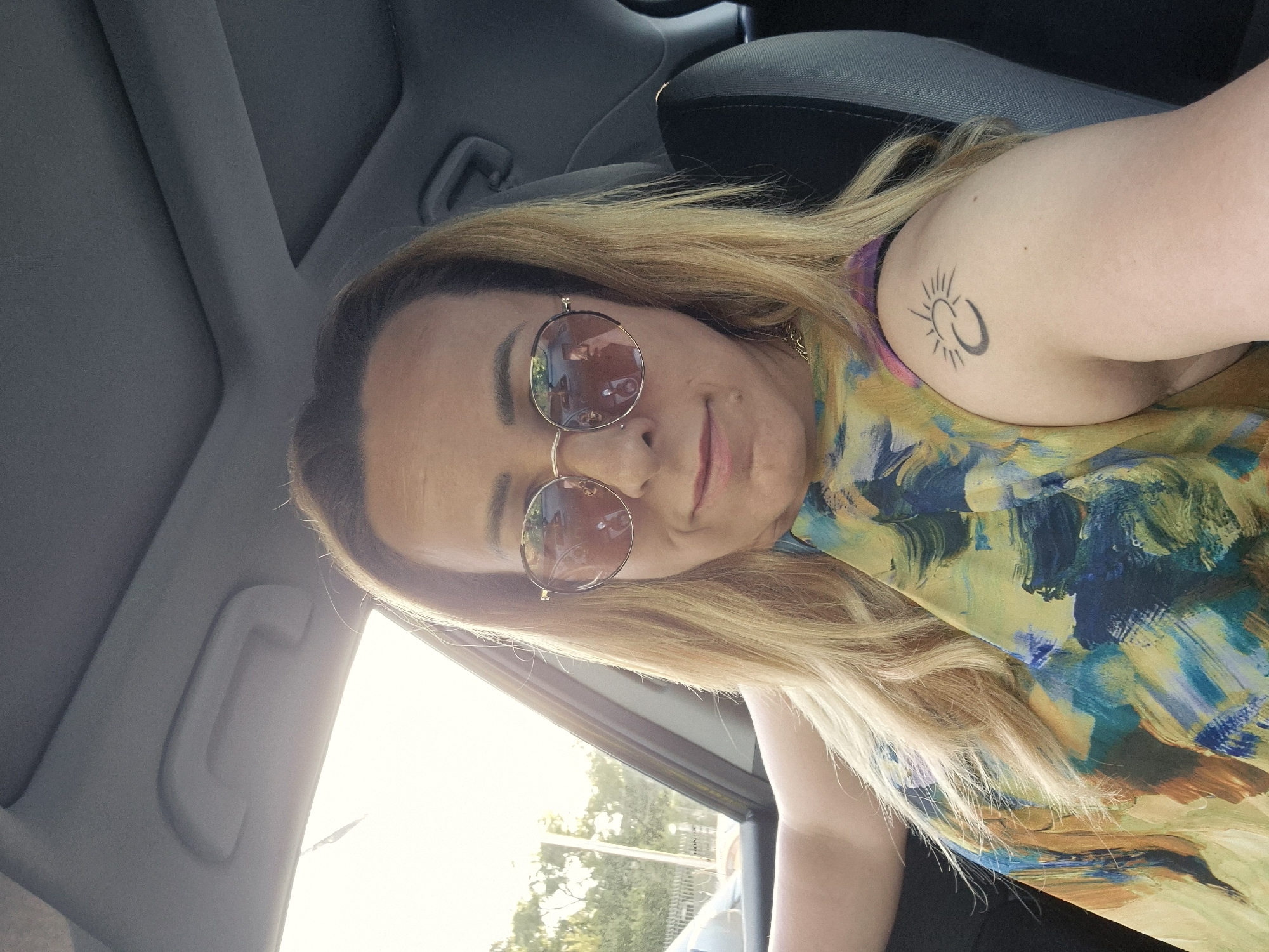 A medium shot photo shows a person with blonde hair and sunglasses sitting in the driver's seat of a car. The person is wearing a colorful, abstract-patterned top. A tattoo of a sun and crescent moon is visible on their upper left arm. The background is blurry, but shows trees and a fence through the car window. The interior of the car, including the headliner and seat, is also visible. The lighting suggests it is daytime. The specific location in the city of Unknown, Unknown is not determinable from the image.
