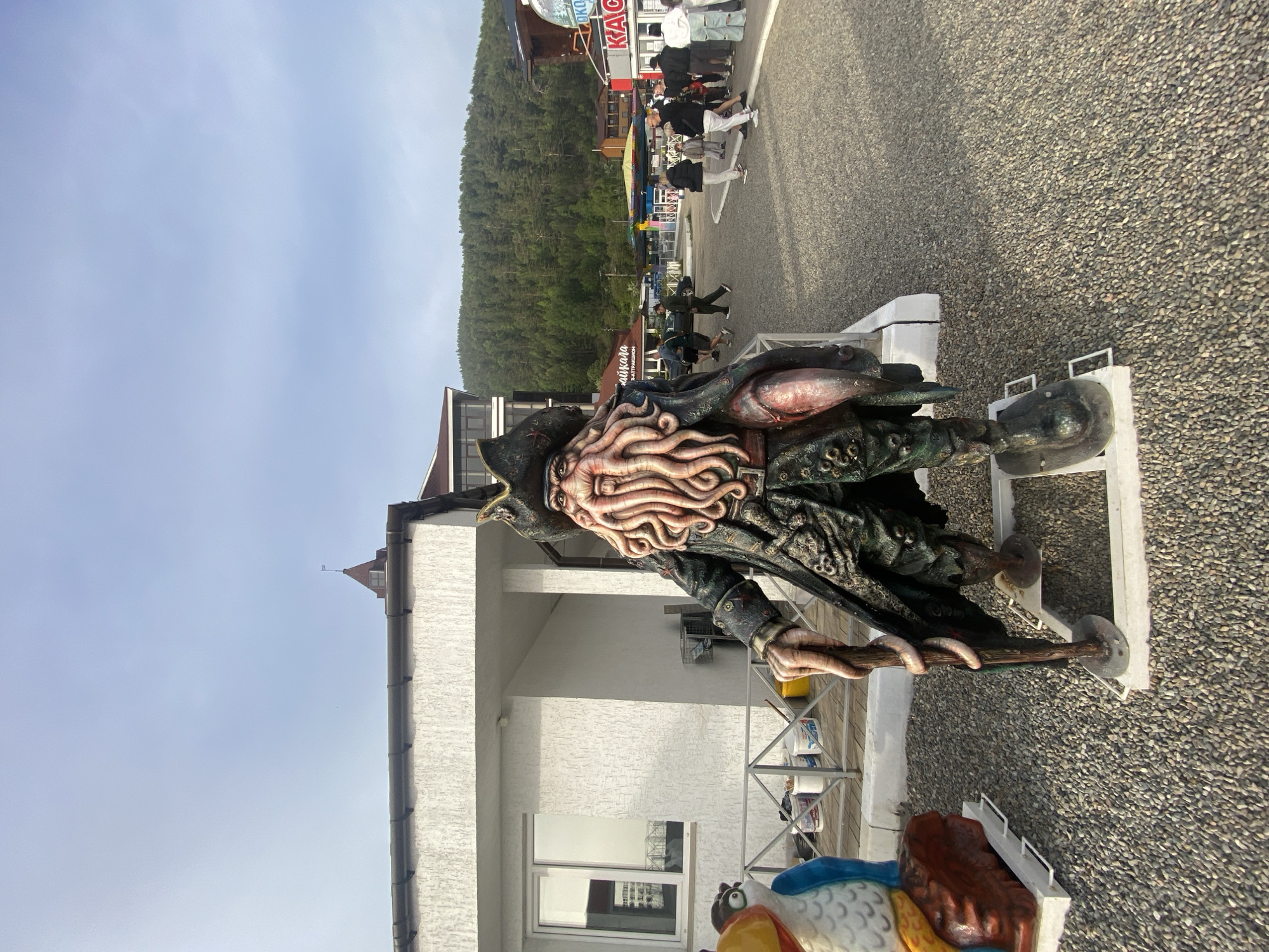 In Listvyanka, Russia, a life-size statue resembling Davy Jones from Pirates of the Caribbean stands in the foreground. The statue is detailed with tentacles for a beard and a pirate hat. To the left of the statue is a colorful, cartoonish bird statue. Behind them, a white building with a window and a railing can be seen. The ground is covered in small gravel. In the background, a paved area leads to various buildings, including a red structure with the word "KAC" visible, and a forested hill rises behind the buildings under a pale blue sky. Several people are walking in the background.