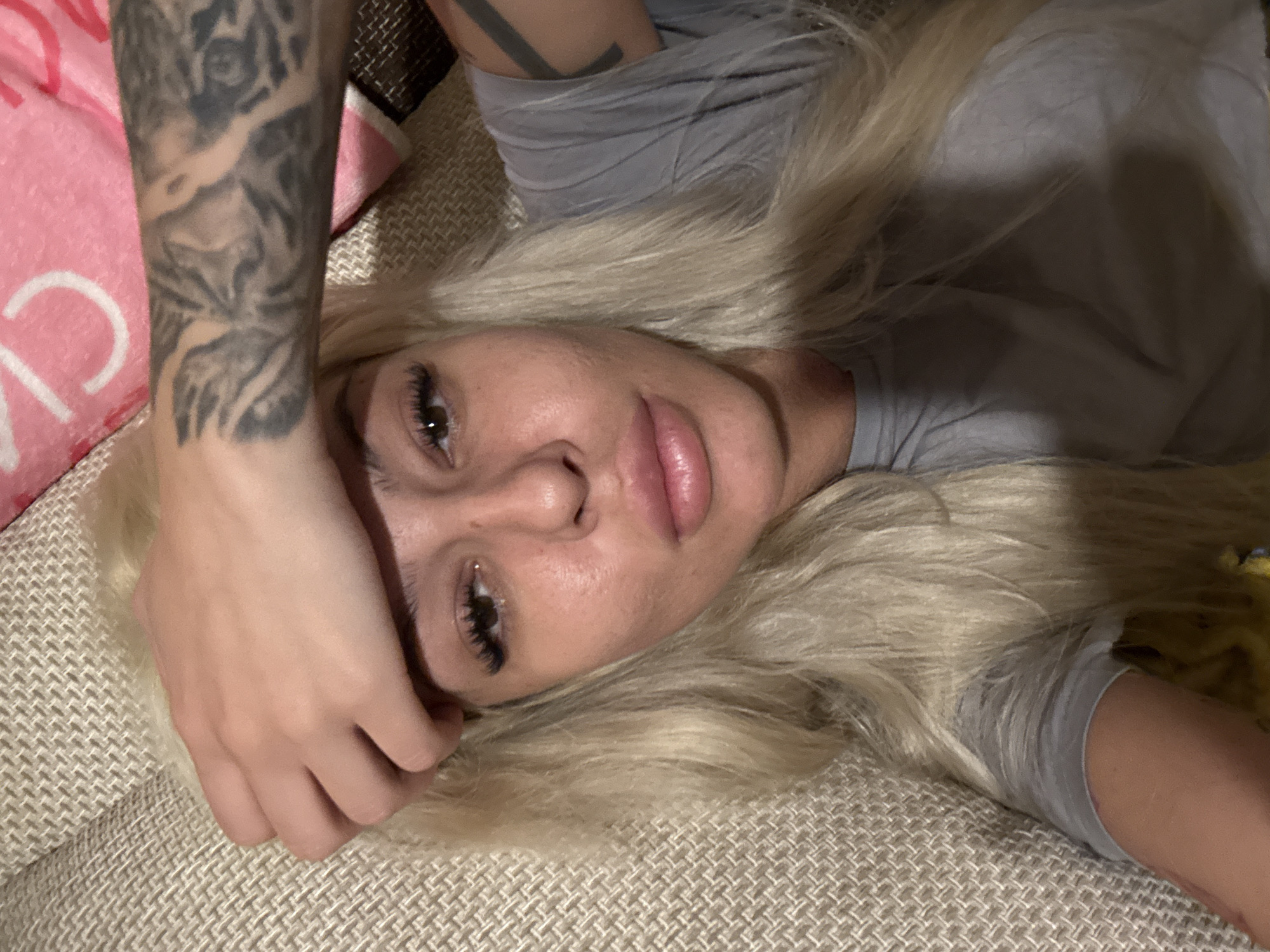 A woman with platinum blonde hair lies on a textured beige surface, potentially a couch or bed. She is wearing a gray t-shirt. The woman's head is slightly tilted, and she looks directly at the camera with a soft expression. She has noticeable eyelash extensions and full lips. Her left arm is raised, and her hand is resting near her forehead. A full-sleeve tattoo is visible on her left arm. In the upper left corner, a pink and white object is partially visible, possibly a blanket or pillow. The location is unknown.