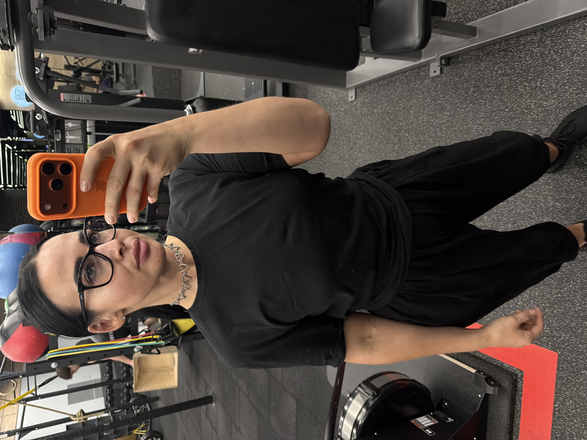 A woman is taking a selfie in what appears to be a gym. She is wearing black framed glasses, a black shirt, and black pants. She is holding an orange smartphone to take the picture. Exercise equipment is visible in the background, including machines, weights, and resistance bands. There is no indication of Saint Petersburg, Russia in the image.