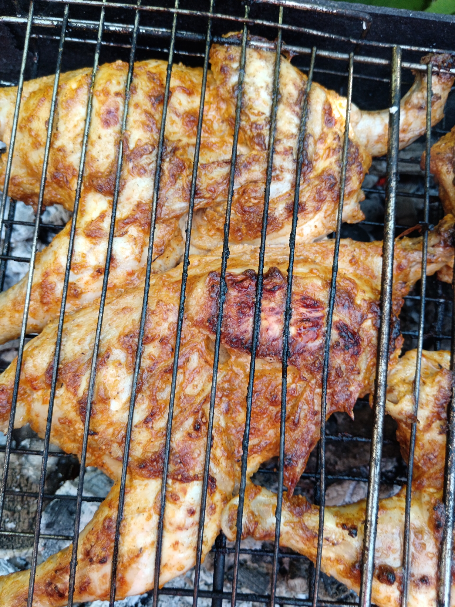 A close-up, overhead shot shows chicken pieces grilling on a black metal grate over coals. The chicken is seasoned and appears to be well-cooked, with some char marks from the grill. The scene is an outdoor setting. The image does not provide specific details about the location in the city of Sorochinsk, Russia, or the presence of any people.