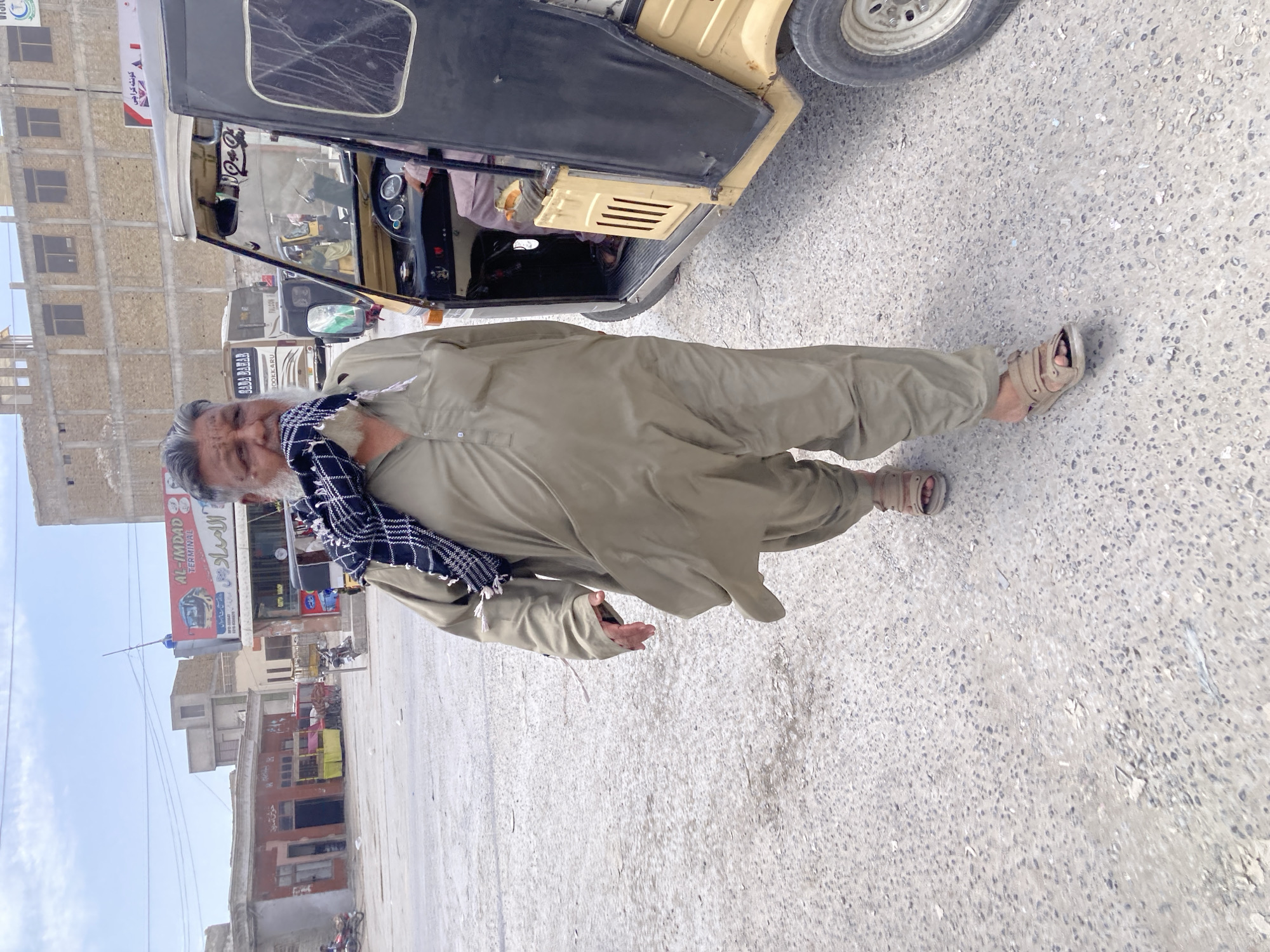 An outdoor, eye-level shot captures a smiling man standing on a paved street next to a three-wheeled auto-rickshaw (tuk-tuk) under bright daylight. The man, likely middle-aged or older, has a grey beard and is wearing a light green traditional shalwar kameez and a black and white checkered scarf around his neck. He is looking directly at the camera, with one hand slightly extended.
Behind him, a beige and black auto-rickshaw is parked, with a driver partially visible inside. Further back, along the street, are various buildings, some appearing residential and others commercial, suggesting an urban or semi-urban setting. The sky is clear, indicating good weather.
Visible text in the background includes:
* A red sign that reads "AL-IMDAD TERMINAL" and "الامدادی" (Al-Imdadi) in Urdu.
* A white banner with Urdu text "سویٹ کی خوشبو کراچی" (Sweet Ki Khushboo Karachi - "The scent of Sweet Karachi").
* On a vehicle visible behind the auto-rickshaw: "SADA BAHAR" and "POK-KARU".
* On a small sign above a shop: "AUTO SHOP".
The ground is a light-colored, gravelly pavement. The overall scene depicts a bustling street in what appears to be Quetta City Tehsil, Pakistan.
