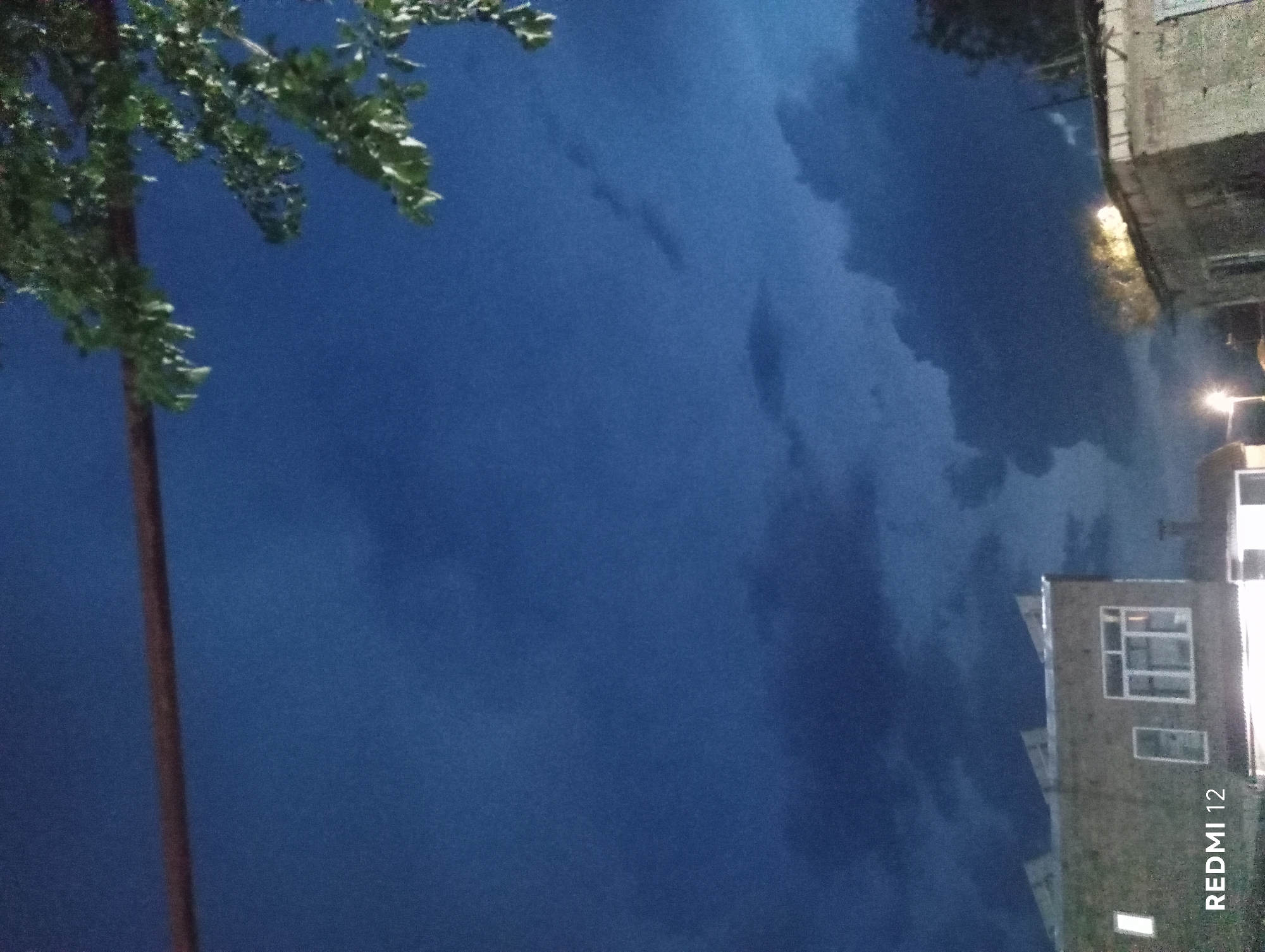 A night-time, low-angle shot captures a dark blue sky with scattered, ominous clouds. In the foreground, the tops of two buildings are visible, one on the left with a large window and the other on the right. A horizontal dark object, possibly a wire or pole, runs across the upper part of the frame, with green leaves from a tree hanging from the right side. The word "REDMI 12" is overlaid on the lower left corner of the image.