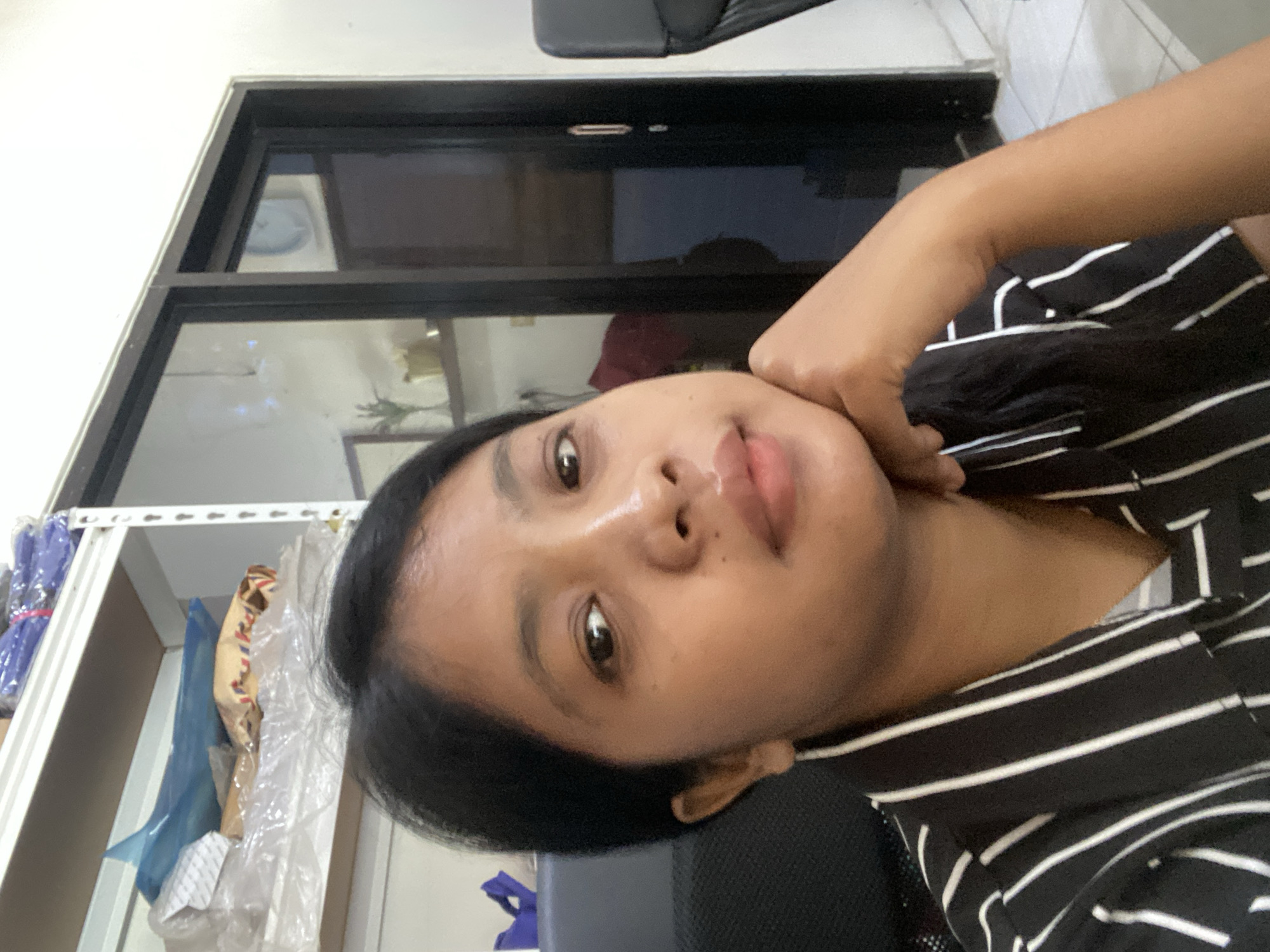 A person with dark hair and brown skin is visible in this indoor selfie. They are wearing a black and white striped shirt. The background features a dark-framed window and a shelf with items wrapped in plastic. Based on the location provided, the photo was likely taken in Negara, Indonesia.