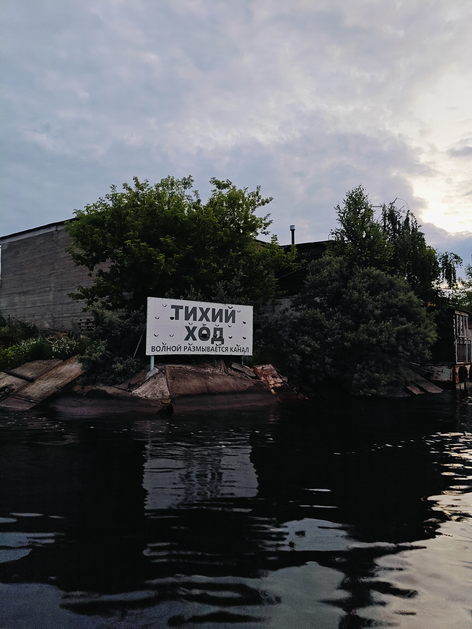 The image shows a sign in Russian that reads "ТИХИЙ ХОД" (Tikhij Khod), which translates to "Slow Speed" or "Gentle Movement." Below this, it says "ВОЛНОЙ РАЗМЫВАЕТСЯ КАНАЛ" (Volnoy razmyvaetsya kanal), meaning "The channel is being eroded by waves." The sign is white with black text and is positioned in front of lush green trees and a large, somewhat dilapidated concrete building. The scene appears to be near a body of water, likely a canal, given the text on the sign. The water is dark and reflects the sky and surrounding elements. The sky is overcast with hints of sunlight breaking through the clouds. The overall impression is one of an industrial or semi-abandoned waterfront area in Chaikovsky, Russia, warning about the potential for erosion caused by boat traffic. There are no people visible in the image.