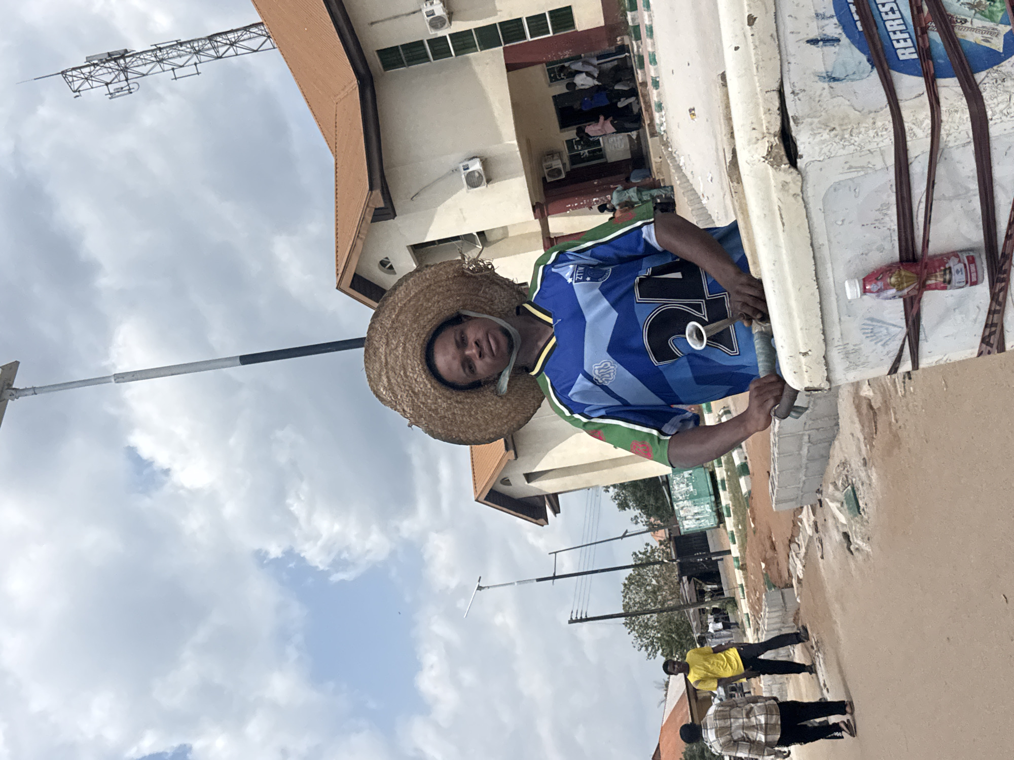 An outdoor scene in Ihiagwa, Nigeria, captured during daylight under a partly cloudy sky. The foreground is dominated by a dark-complexioned male wearing a straw hat and a blue and green "ZTIV 24" sports jersey, with a blue face mask around his neck. He stands behind a white, weathered cooler, holding a white handle protruding from its top. A red-patterned bottle is strapped to the side of the cooler, which also displays remnants of a "REFRESH" label.
The ground consists of a dirt or unpaved road with sections of concrete blocks and loose soil indicating construction or disrepair. Green and white painted curbs line parts of the roadside. To the left, two men walk away from the camera: one in a plaid shirt and the other in a yellow t-shirt. In the mid-ground, a tall streetlight with a solar panel is visible, alongside other utility poles.
The background features a two-story building with a reddish-brown tiled roof, light-colored walls, and a prominent entrance. Several individuals are gathered near this entrance. Multiple air conditioning units are affixed to the building's facade. To the far right, a tall, metal lattice telecommunications tower with antennas rises above the roofline. Additional residential or commercial structures with reddish roofs are partially visible in the distant left background.