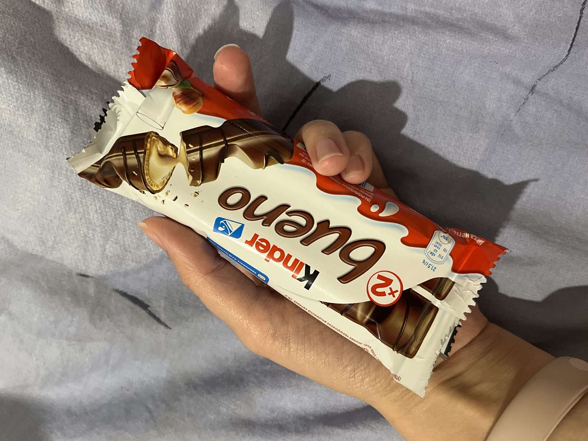 The image depicts a hand holding a Kinder Bueno chocolate bar on a grey surface. The chocolate bar's packaging is partially torn open at the top, revealing the chocolate inside. The packaging is white with a brown "Kinder Bueno" logo and an image of the chocolate with hazelnuts. The background is a grey, slightly textured surface. The photo was not taken in Verkhnyaya Pyshma, Russia.