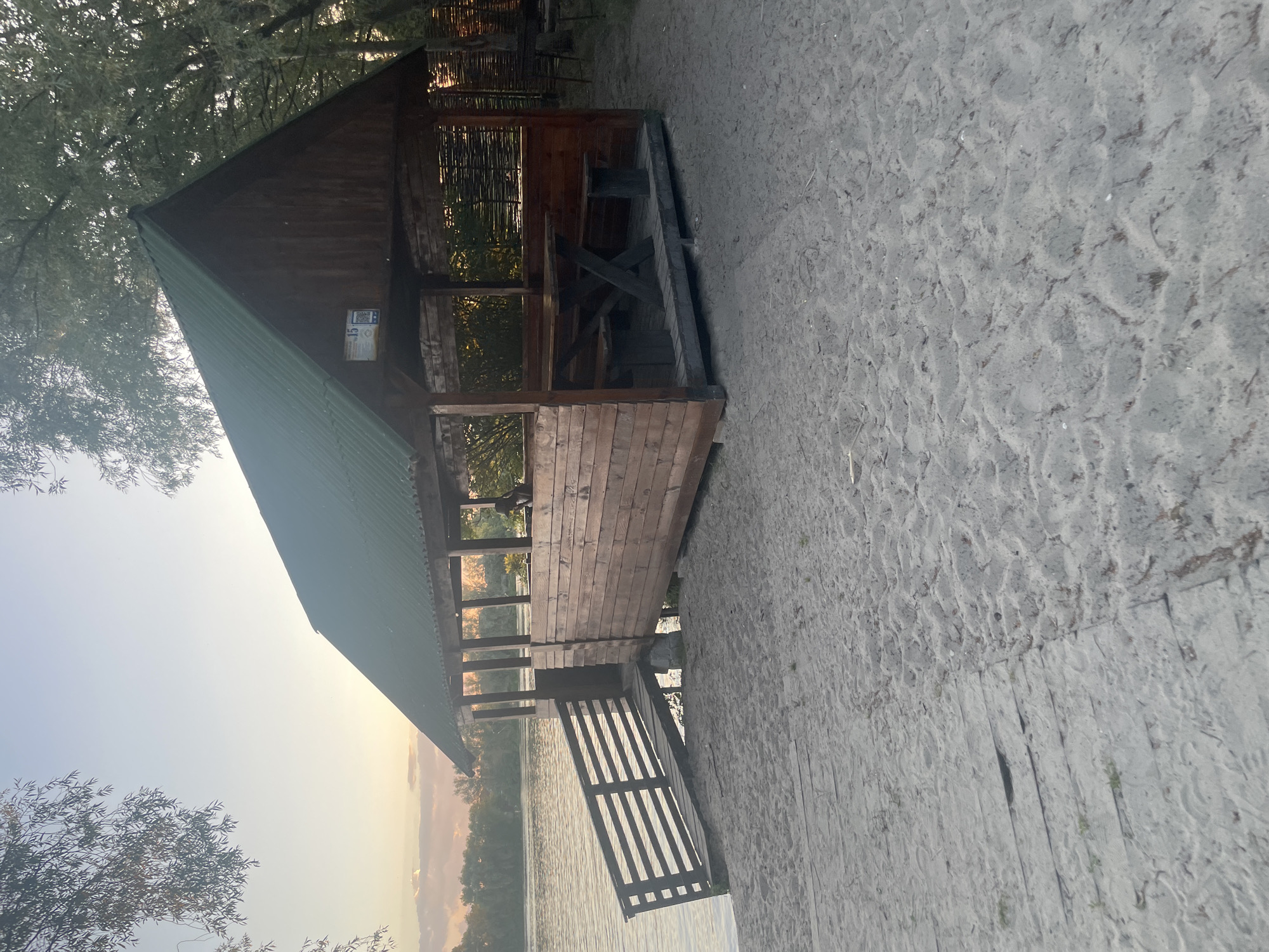This media file depicts a wooden gazebo with a green metal roof situated on a sandy area next to a body of water, likely a river or lake. The gazebo appears to be open-sided with wooden walls and a rustic fence. Inside, a wooden table and benches are visible. A wooden staircase leads from the gazebo down towards the water. The scene is bathed in the soft light of either sunrise or sunset, casting a warm glow and long shadows. The sky is clear with hints of pale yellow and pink hues, suggesting fair weather. Trees with green foliage surround the gazebo, some branches reaching towards the structure. The sandy ground shows imprints, possibly from footsteps.
The image is oriented vertically. The primary subjects are the gazebo and its immediate surroundings, including the water, sand, and vegetation. There are no people or animals clearly visible in the frame. The overall mood is serene and tranquil, evoking a sense of peace and natural beauty.
On the wooden wall of the gazebo, a rectangular sign with some text and a QR code is attached. The text on the sign is not legible in the provided image. The location context is Kyiv, Ukraine.