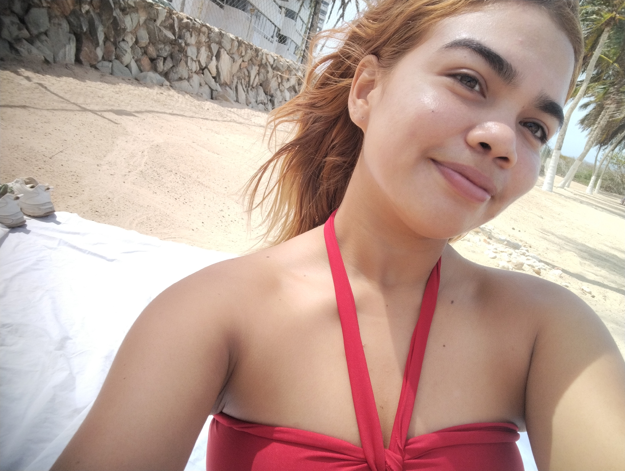 The image is a selfie of a light-skinned woman with reddish-brown hair, wearing a red top. She is on a beach, with sand visible in the background. Behind her, there is a stone wall and some vegetation including palm trees. A white sheet is laid on the sand near her. The presence of the palm trees may indicate that it is likely on the beach in the city of Porlamar, Venezuela. There is a pair of white shoes beside the white sheet.