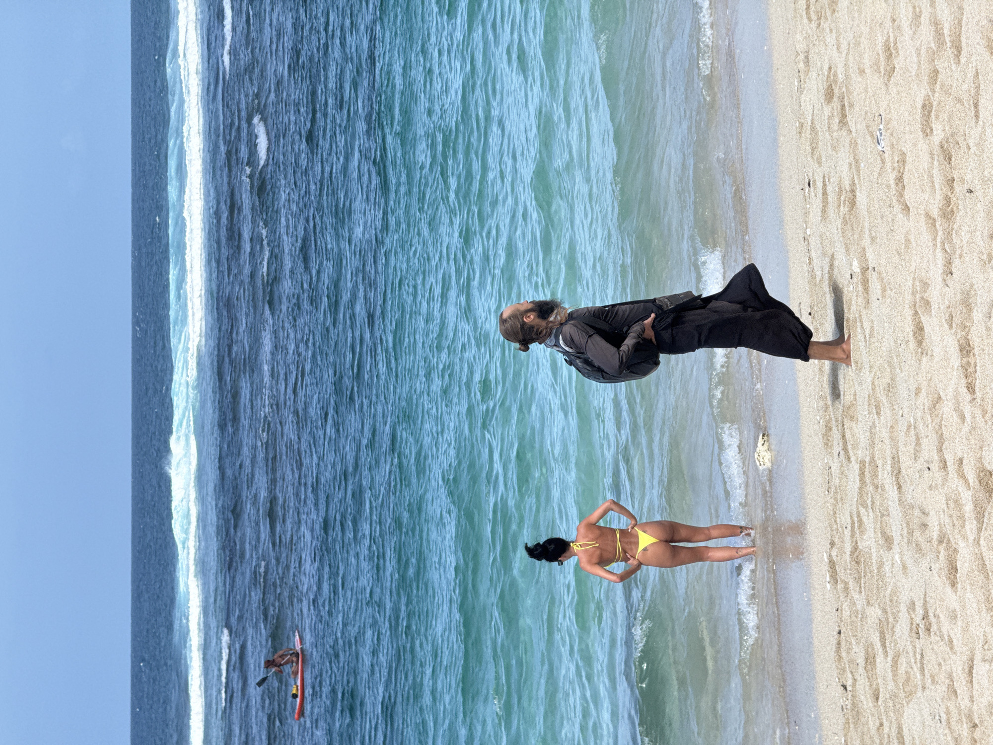 The image depicts a beach scene in Kuta Selatan, Indonesia. The horizon is visible on the left side of the image, with the sea occupying the majority of the background. A surfer is visible in the ocean. A woman wearing a yellow bikini stands at the edge of the water. To her right is a man wearing a black outfit, with a backpack. He is looking up. The foreground consists of a sandy beach. The image has been rotated 90 degrees clockwise.