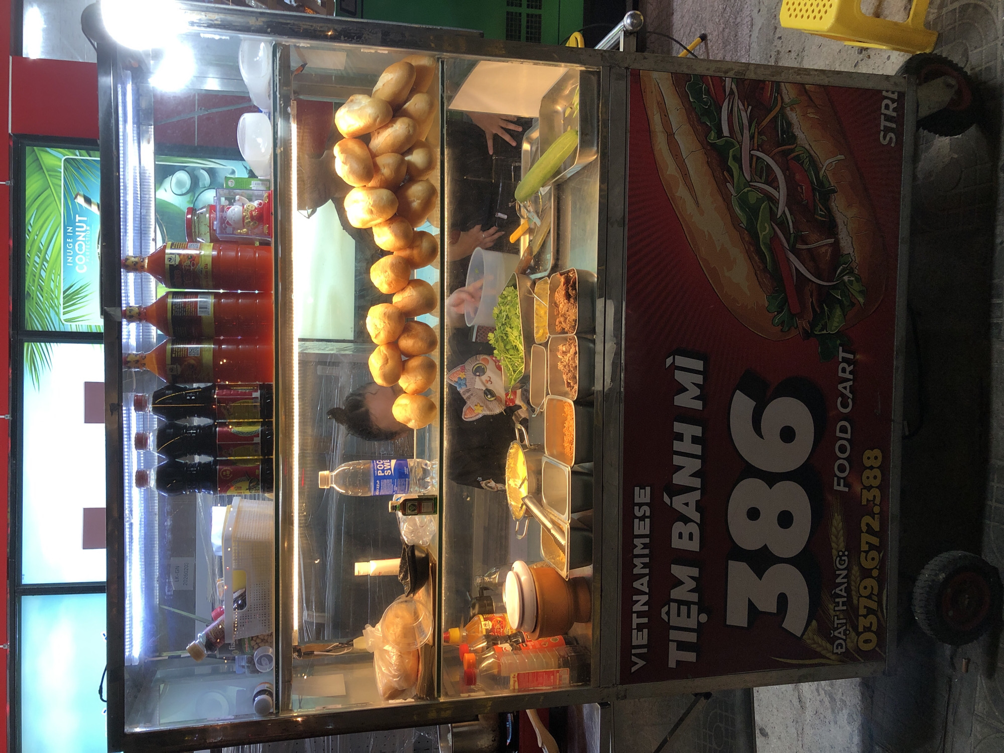 This image depicts a mobile food cart, likely a Banh Mi stand, situated in Đà Nẵng, Vietnam. The cart is a self-contained unit with a glass display case showcasing various ingredients and pre-made items. On the top shelf, rows of bottled sauces and condiments are visible. The middle shelf holds an assortment of fresh ingredients including green leafy vegetables, what appears to be meat fillings, and a long cucumber. There are also several loaves of bread stacked on this shelf. The front of the cart features a large, prominent graphic of a Banh Mi sandwich and text that reads "VIETNAMESE TIỆM BÁNH MÌ 386 FOOD CART" followed by a phone number "0379.672.388". Two wheels are visible at the bottom, indicating its mobile nature. The background suggests an urban environment, with a sidewalk and a yellow plastic stool visible to the right. The lighting is artificial, emanating from within the cart, creating a bright and inviting display. A person, partially obscured, appears to be working behind the counter.