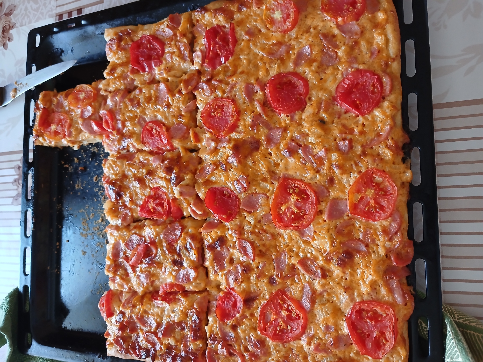 An overhead, close-up shot captures a rectangular pizza on a black baking sheet, set on a table covered with a striped tablecloth. The pizza is cut into squares and topped with melted cheese, sliced red tomatoes, and circular pieces of sausage or pepperoni. A knife with a silver blade and a brown handle rests on the edge of the baking sheet, angled away from the pizza. The pizza appears to be freshly baked, with some crust browning. The lighting is natural and casts soft shadows. The environment suggests an indoor setting, likely a kitchen or dining area. The overall scene is focused on the appetizing appearance of the pizza.