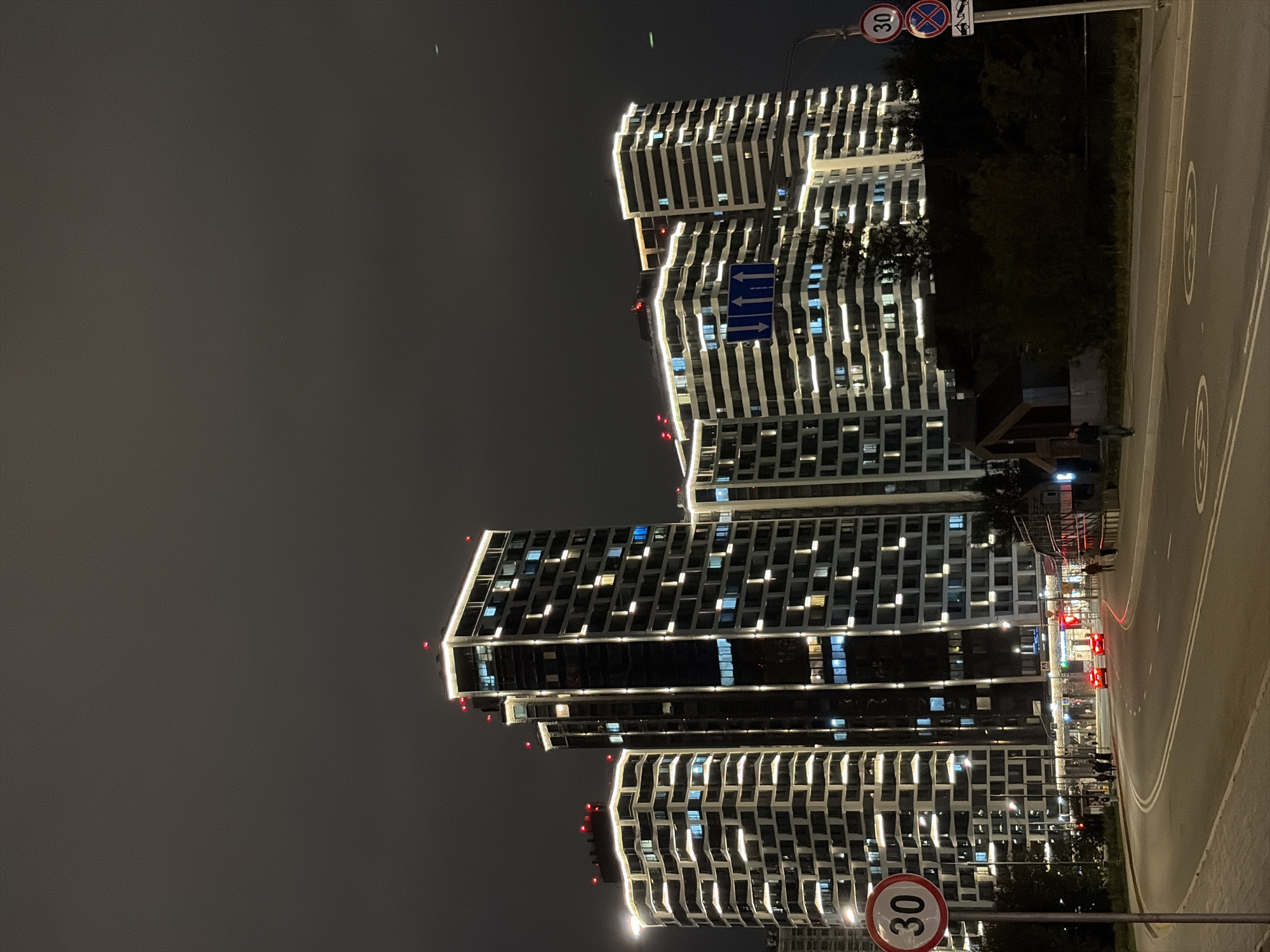 The image captures a nighttime view of the residential complex "Domodedovo Parks" in the city of Moscow, Russia. Several tall, modern buildings with illuminated windows and decorative lighting along their edges dominate the scene. The sky is dark and overcast. In the foreground, a road curves to the left, with a speed limit sign indicating "30" and another sign with a red circle and a diagonal bar over a parking symbol. Red traffic lights are visible in the distance, and a few cars with illuminated headlights are driving on the road. Several pedestrians are walking on the sidewalk. Trees and some lower structures are visible between the road and the residential buildings. The overall impression is one of a contemporary urban environment at night.