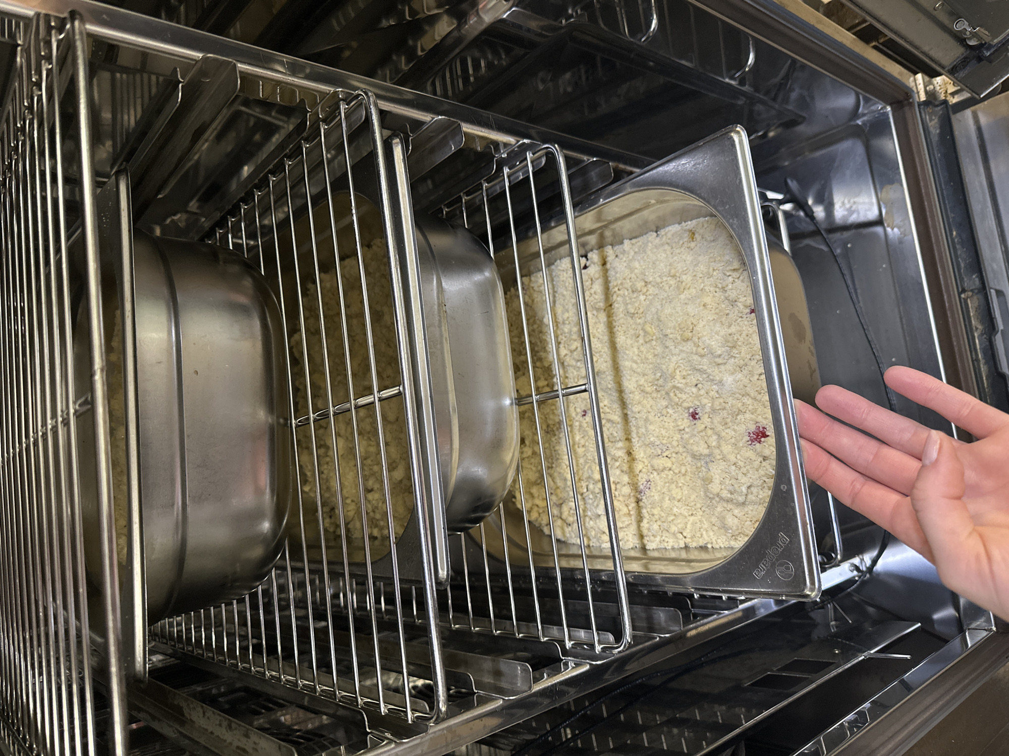 An overhead shot shows a commercial oven with three levels of metal racks holding food. On the top rack, a metal pan holds a beige crumb topping. Below this, a second metal pan contains a similar topping. The bottom rack holds another metal pan filled with the beige crumb topping, which has visible red specks. A person's left hand, with fingernails painted a light pink, is visible in the lower right corner, reaching towards the bottom pan. The oven interior is stainless steel, and the racks are also metal. The scene appears to be indoors, in a commercial kitchen.