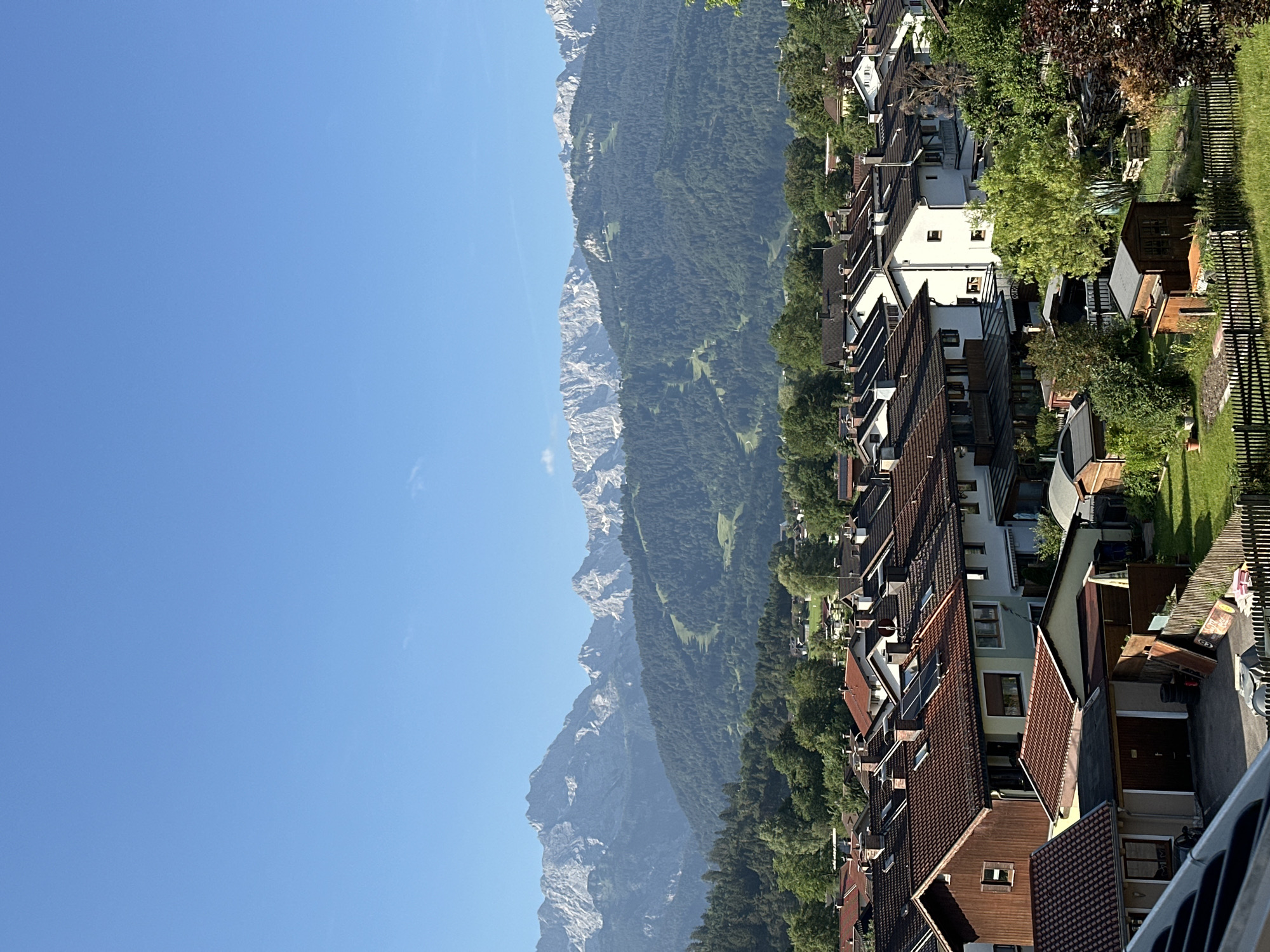 This media file shows a residential area with houses nestled at the foot of a mountain range. The scene is characterized by a clear blue sky, suggesting a bright, sunny day. The houses feature a variety of roof styles, predominantly terracotta tiles, and are interspersed with lush green trees. In the background, the imposing mountains rise, their rocky peaks visible against the sky, partially covered in dense forest. The perspective suggests the photo was taken from an elevated position, looking down on the town and across towards the mountains.
The activity depicted is simply the peaceful existence of a residential area against a dramatic natural backdrop. There are no people or specific events visible, making it a serene landscape shot. Notable details include the texture of the mountain rock, the varied architectural styles of the houses, and the vibrant green of the trees and lawns. The clear sky and bright sunlight indicate it is likely daytime, possibly late morning or afternoon. The location context of Garmisch-Partenkirchen, Germany, aligns with the mountainous landscape and architectural style. There is no visible text in the image.