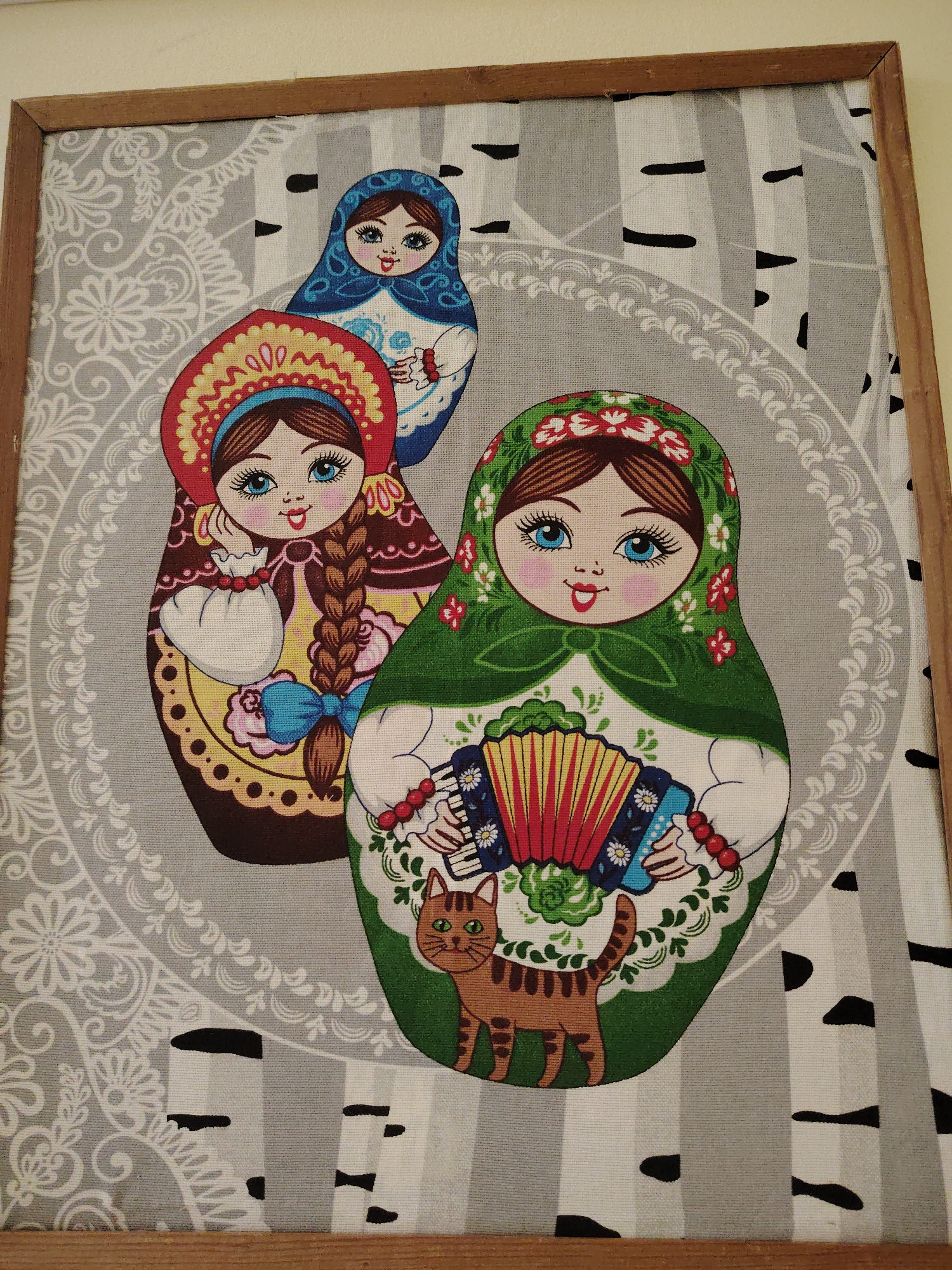 This media file is a framed piece of textile art, likely a fabric print or tapestry, depicting a group of three matryoshka dolls and a cat. The scene has a distinctly Russian folk art aesthetic, fitting the provided location context of Sarapul, Russia.
The primary subjects are three smiling matryoshka dolls, arranged in a layered composition. The largest doll is in the foreground on the right, wearing a green headscarf adorned with red and white flowers, and a white dress with green leaf motifs. It is actively holding and seemingly playing a red and blue accordion. Its eyes are bright blue. A brown striped cat with green eyes stands at the base of this doll.
Behind and to the left of the largest doll is a medium-sized matryoshka. This doll wears a red and gold kokoshnik-style headpiece and a brown dress with yellow patterns. It has a long brown braid tied with a blue bow and holds a white drawstring pouch in its hands. Its eyes are also blue.
The smallest doll is positioned at the top center, behind the other two. It wears a blue headscarf with white patterns and a white dress, holding a small, indistinct white object.
The background features a pattern of grey and white vertical stripes, resembling birch tree trunks with distinctive black markings. A large grey circular frame with intricate white floral and paisley patterns encircles the dolls, separating them from the birch tree background.
The artwork is set within a simple, light brown wooden frame. The overall impression is cheerful and decorative. No visible text is present in the image.