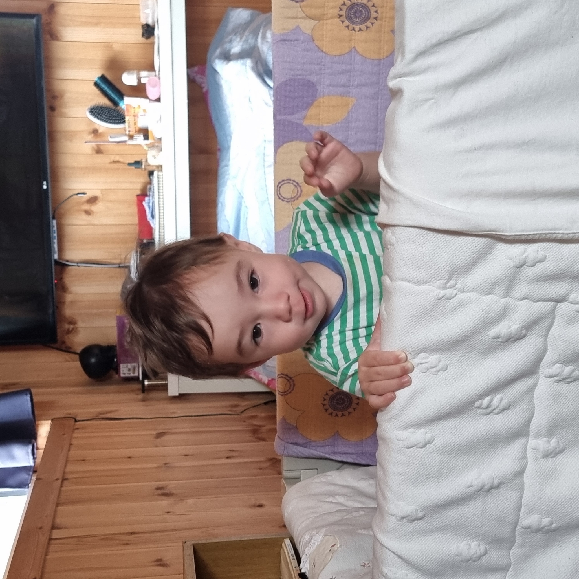 In Dunnae-myeon, South Korea, a young child with brown hair and large, dark eyes is peeking over the edge of a white crib. The child is wearing a green and white striped t-shirt and has their hands resting on the crib's edge. The crib is covered with white bedding that has a textured pattern of small, raised floral shapes. Behind the crib, a colorful blanket with a purple and yellow floral design is visible. The background consists of wood-paneled walls and a shelf with various items, including a television, a black speaker, and grooming accessories.