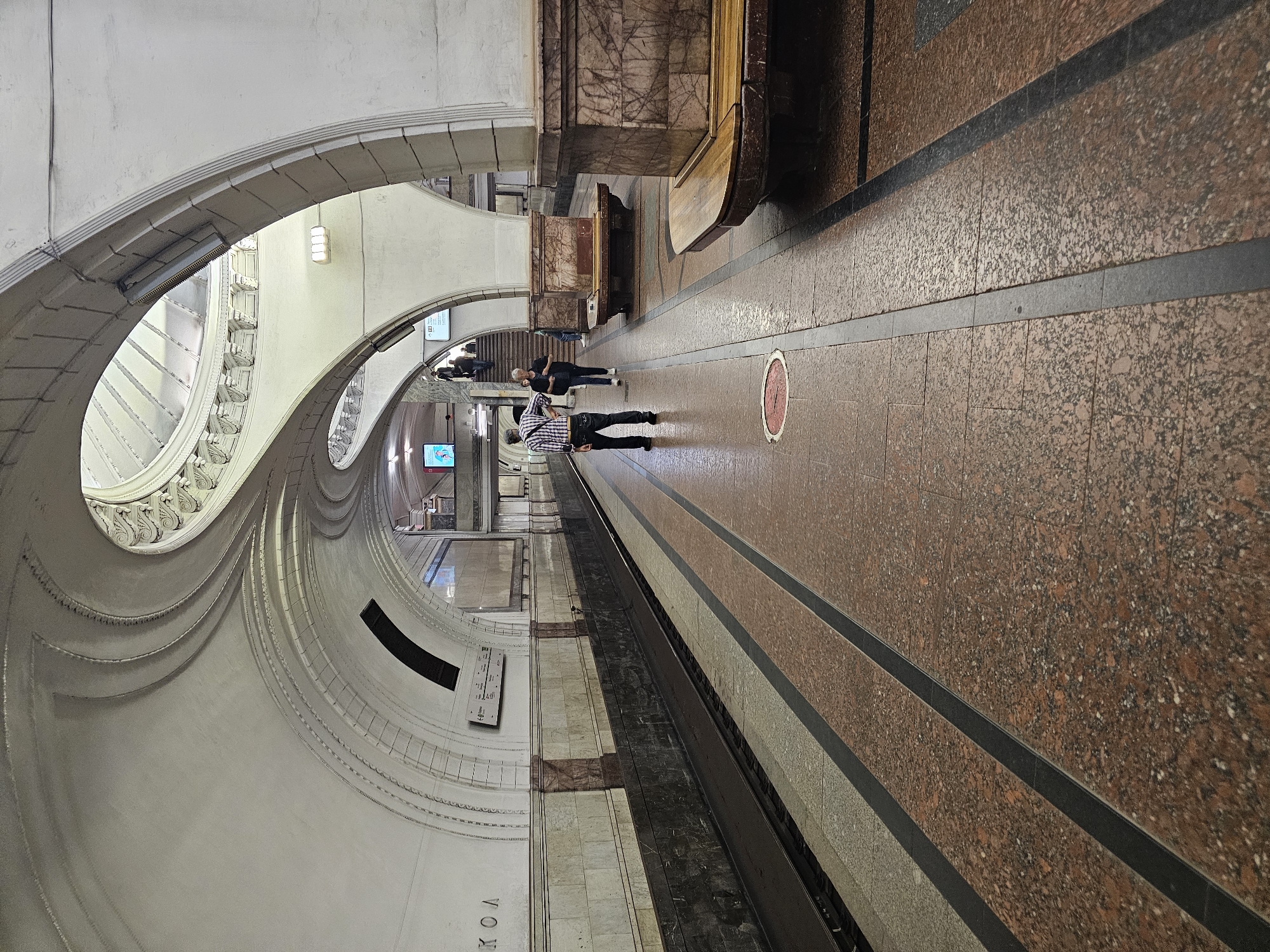 This is a photograph taken inside the Moscow Metro system, specifically at the Mayakovskaya station, a well-known example of Art Deco architecture. The image depicts a subway platform with distinctive vaulted ceilings and ornate architectural details, characteristic of the station's design.
The platform floor is made of polished granite in shades of red and beige, with black strips dividing different sections. Two sets of train tracks are visible, separated by the platform. Along the platform, there are wooden benches with ornate marble supports.
In the mid-ground of the image, two individuals are present. A man in a checkered shirt and dark pants stands facing towards the left, with his hands on his hips. To his right, a woman in dark clothing walks away from the camera, heading towards a staircase in the background. Further in the background, several other people are seen ascending a staircase, and another person is visible on a different platform.
The station's architecture features large, curved arches that create a sense of grandeur. The ceiling is a complex structure of white plasterwork with decorative moldings. The walls are covered with marble panels in various colors, including pink and cream. Signs with Cyrillic text are mounted on the walls, indicating station information and directions. A digital display screen is also visible on the left side of the platform. The lighting in the station appears to be a mix of natural light filtering from above and artificial light fixtures. The overall atmosphere suggests a functional, yet artistically significant, public transportation space.